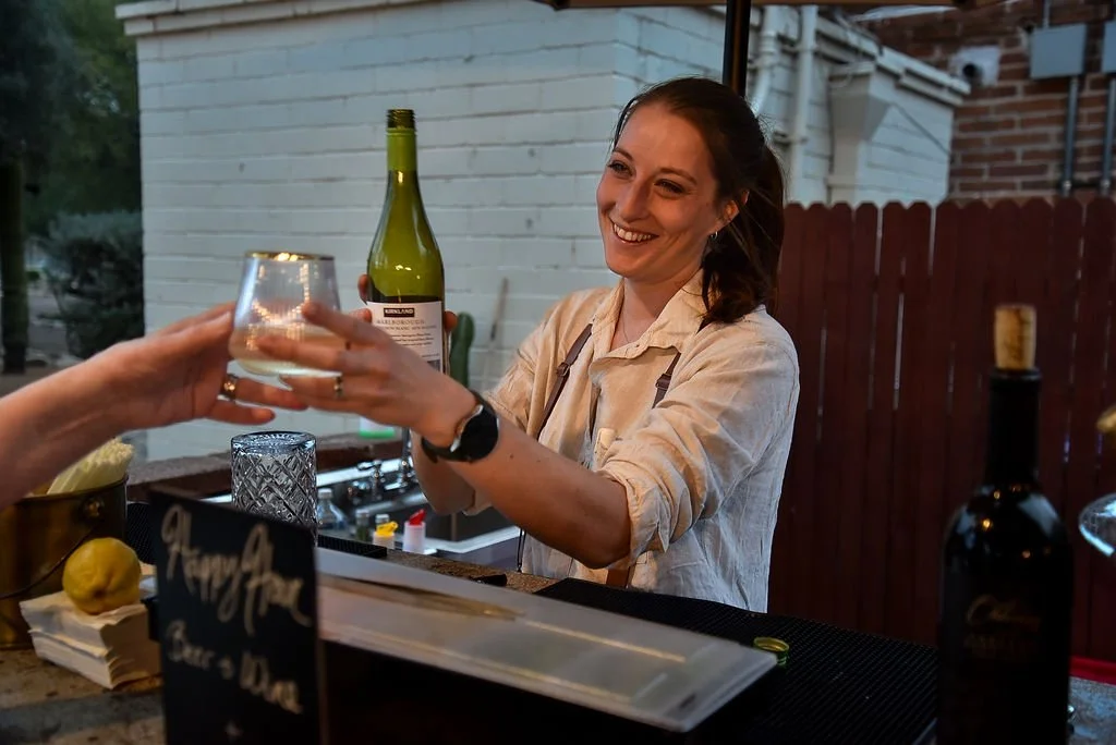A female bartender smiles as she serves a glass of white wine over the counter of an outdoor bar at a wedding.