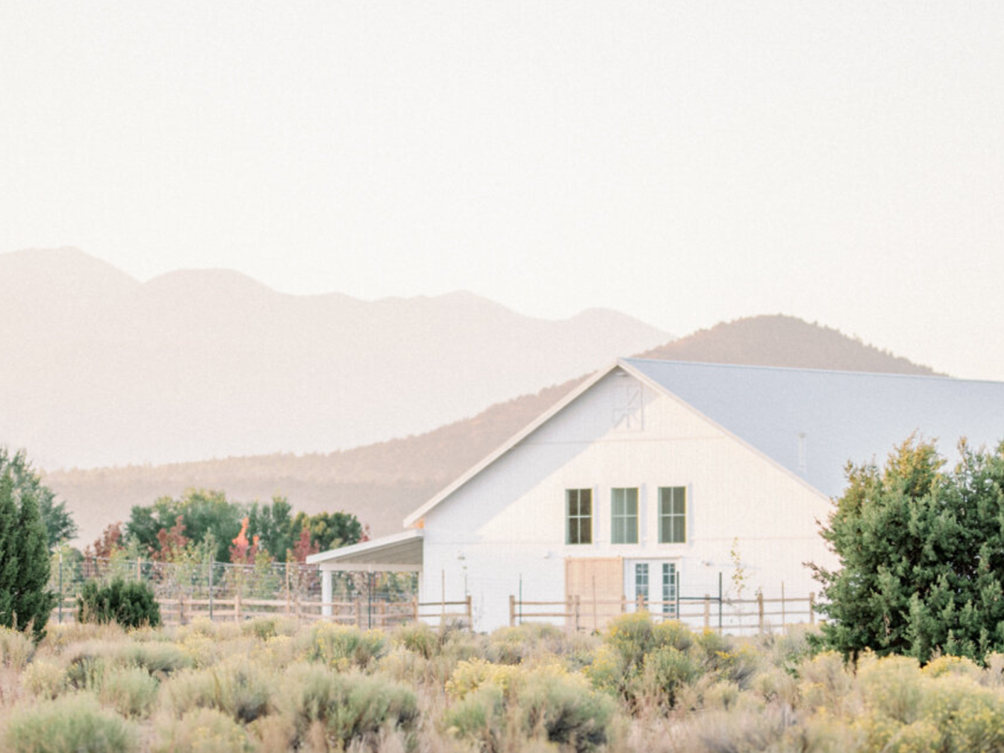 A dreamy white barn sits amongst green grass and yellow bushes, with a backdrop of hazy mountains in Flagstaff.