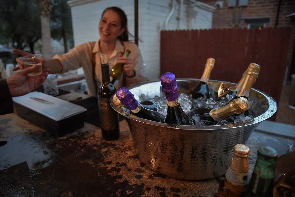 A female bartender smiles as she serves white wine to a guest over the counter of an outdoor bar. In the foreground is a bucket of champagne and white wine on ice.