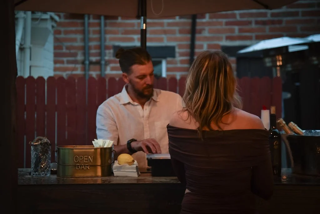 A male bartender in a white linen shirt serves a cocktail to a wedding guest at an outdoor mobile bar.
