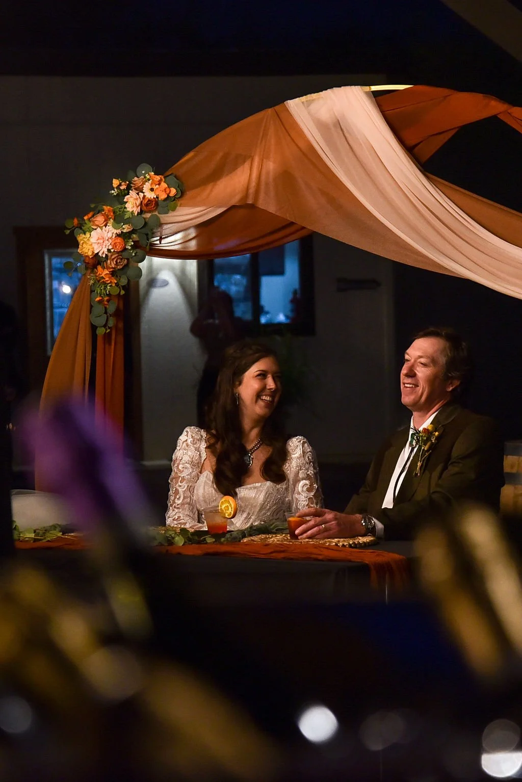A newlywed couple enjoys cocktails at their Arizona wedding. The sit under an arch decorated with orange and white linen and flowers.