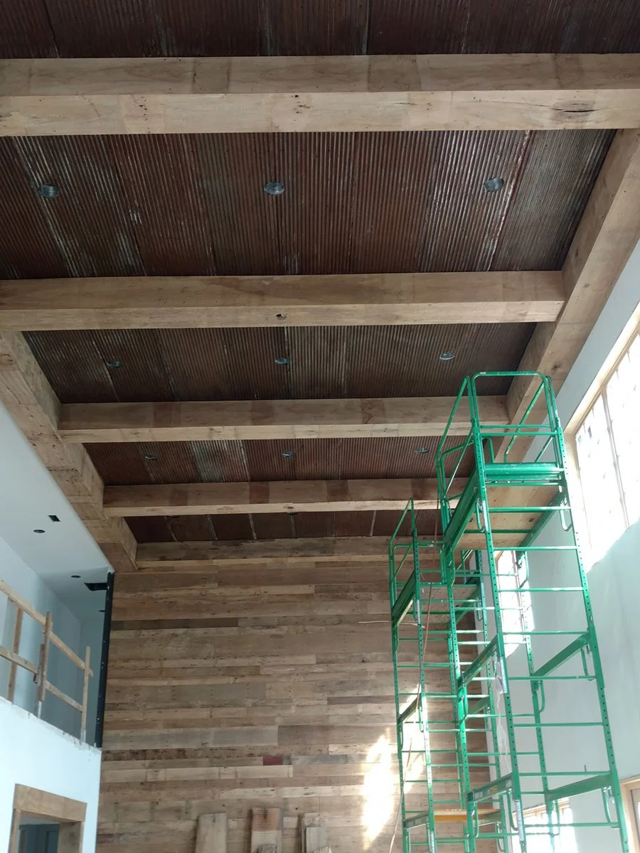 Custom reclaimed barnwood ceiling in great room living area with reclaimed barn wood focal wall or accent wall. Reclaimed rusted barn corrugated roofing material attached to ceiling. Mitered box built ceiling beam detail with a natural finish.