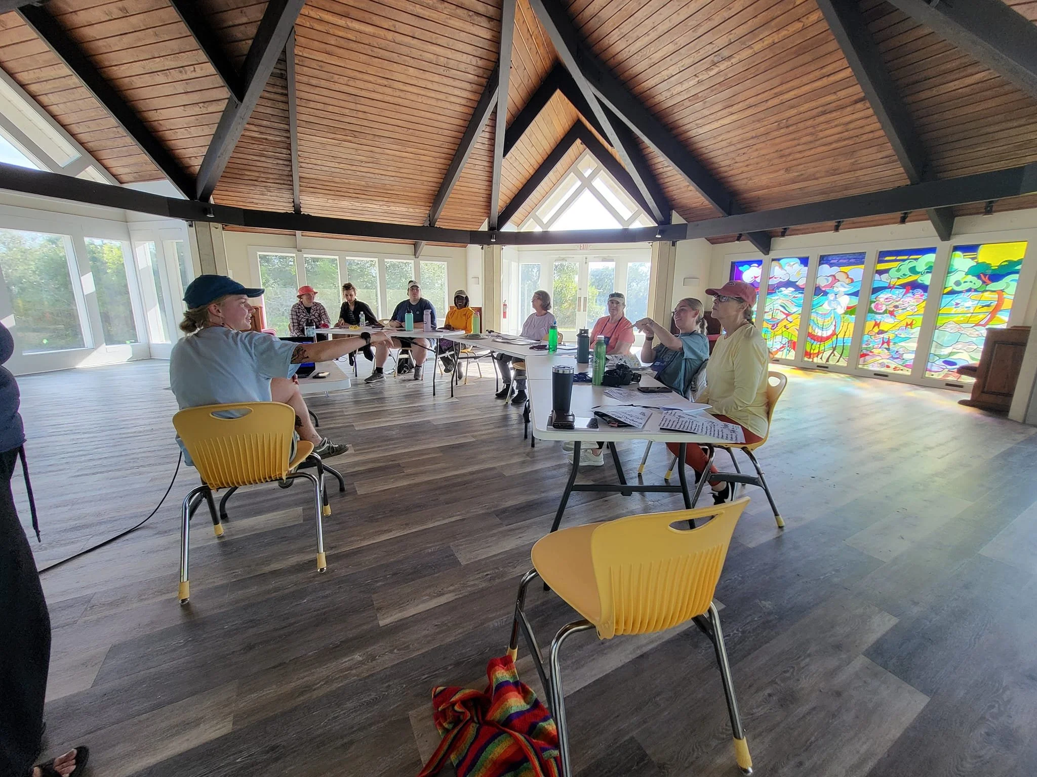 A group of people seated around tables in a room with wooden ceiling beams and large windows, engaging in a discussion or meeting.