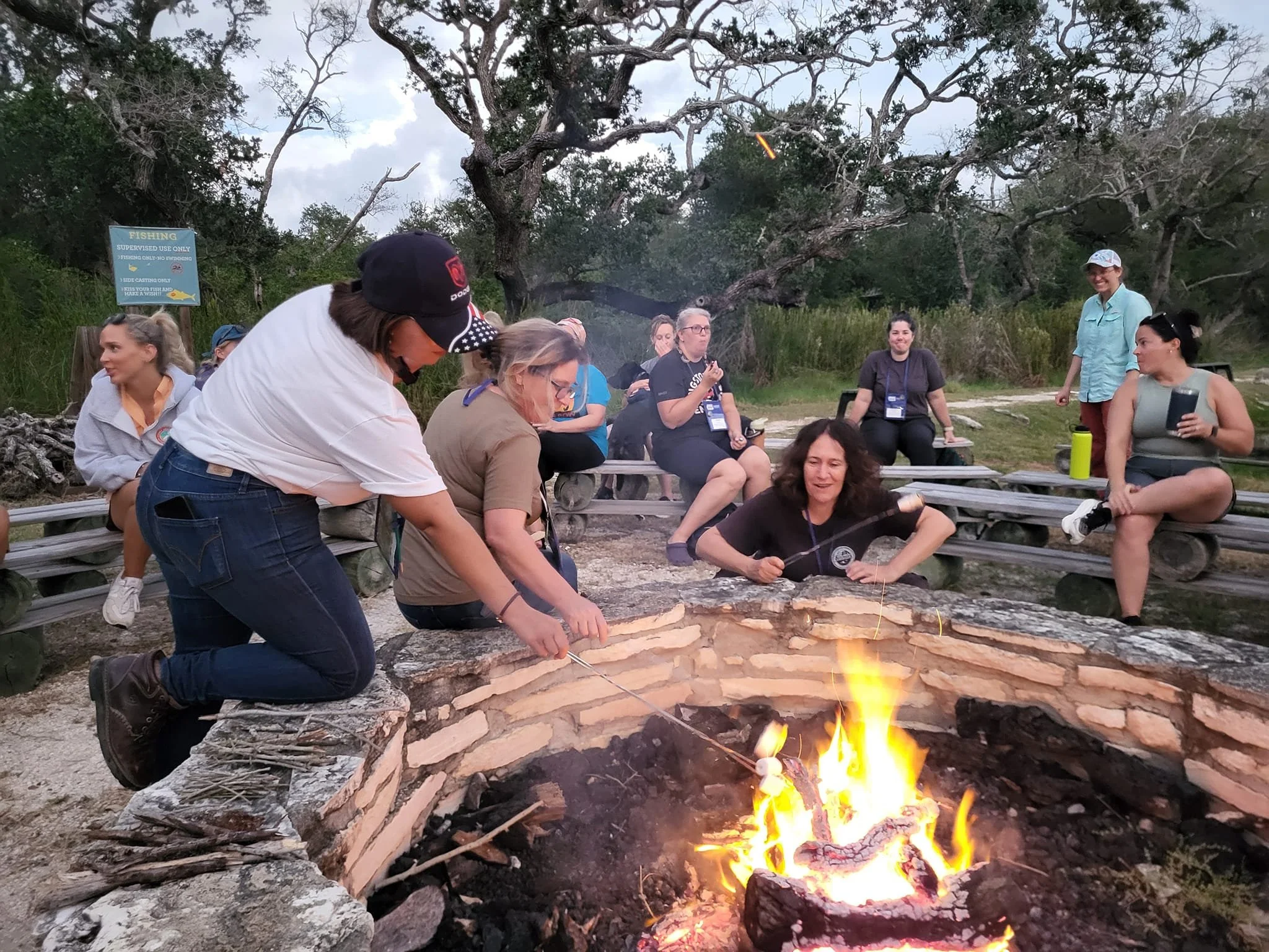 Group of people sitting around a campfire in an outdoor setting, some roasting marshmallows, others sitting and chatting, with trees and a sign in the background.