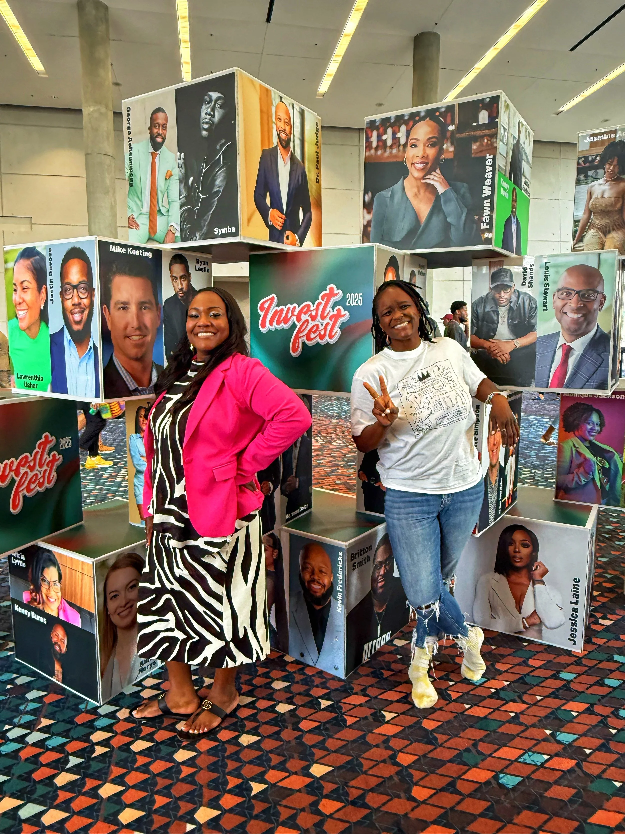 Danicka Brown-Frazier, founder of Alkemi, in a white t-shirt giving a peace sign alongside a fellow entrepreneur in front of the Invest Fest 2025 speaker wall in Atlanta, Georgia.