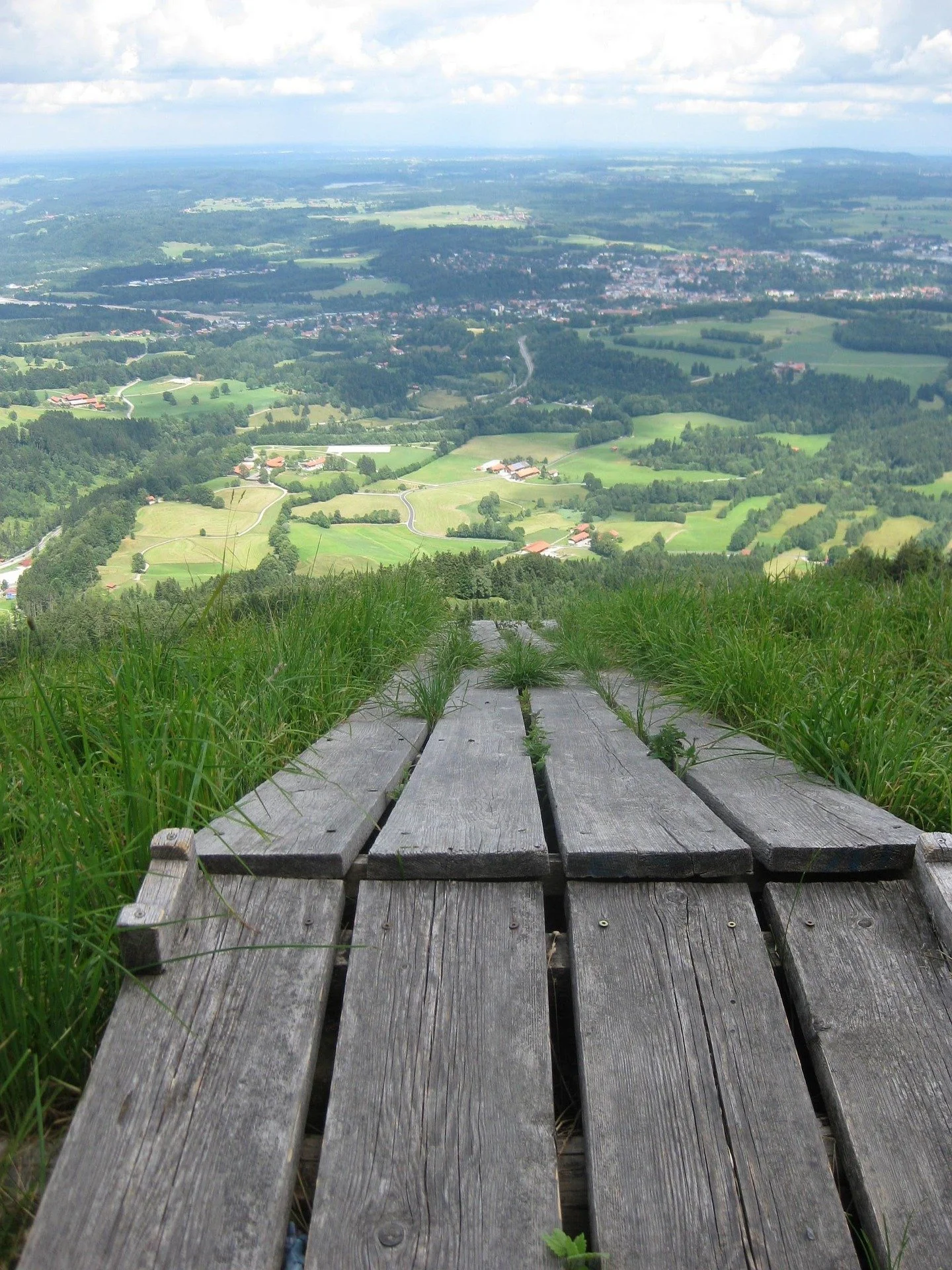 A slatted wooden walkway leads downhill through the green grass toward fields below during the day
