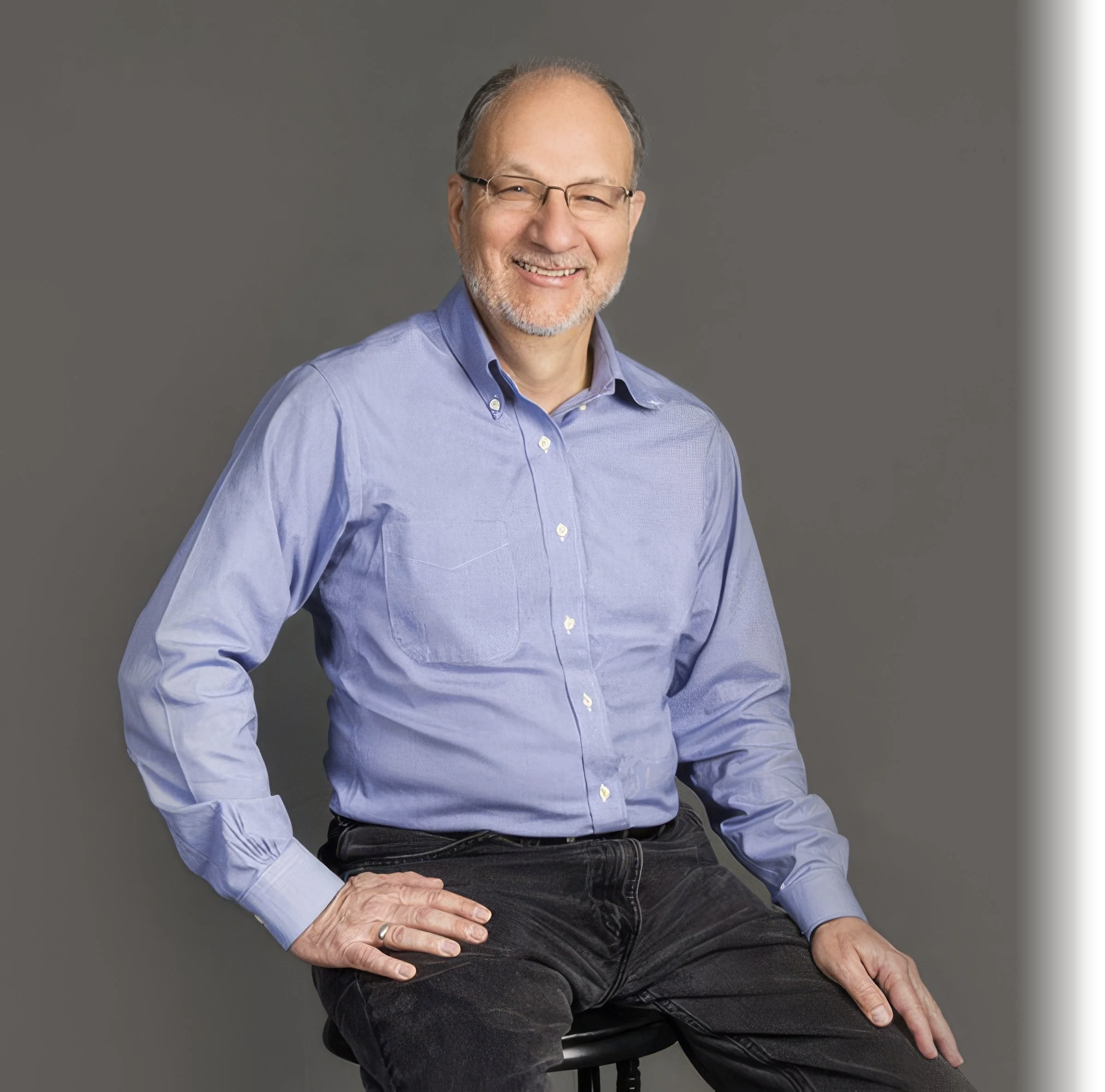 A smiling middle-aged man with glasses and a gray beard, wearing a light blue button-up shirt and black pants, sitting on a stool against a gray background.