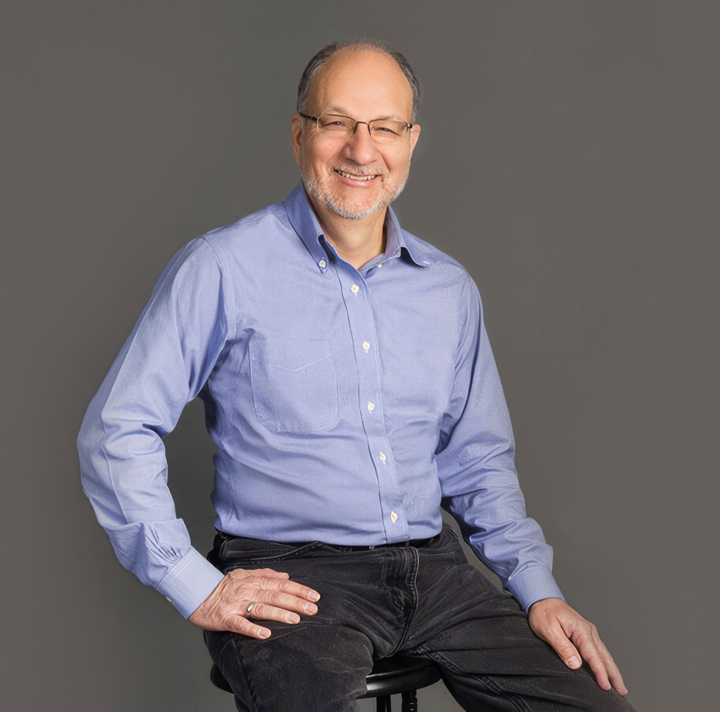 A middle-aged man with glasses and grey hair, wearing a light blue button-up shirt and black pants, sitting on a stool against a grey background, smiling at the camera.