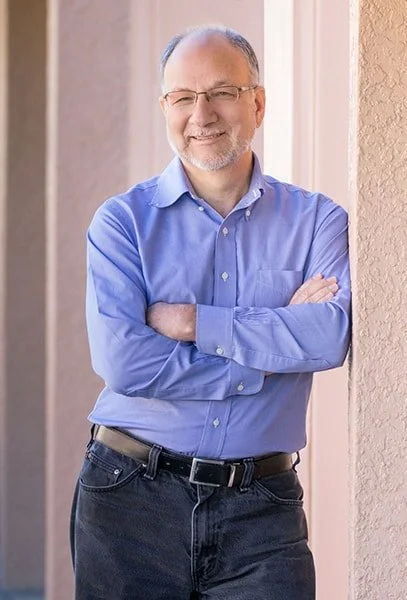 A middle-aged man with glasses and a beard, wearing a blue button-up shirt and dark jeans, smiling with arms crossed, standing by a window.