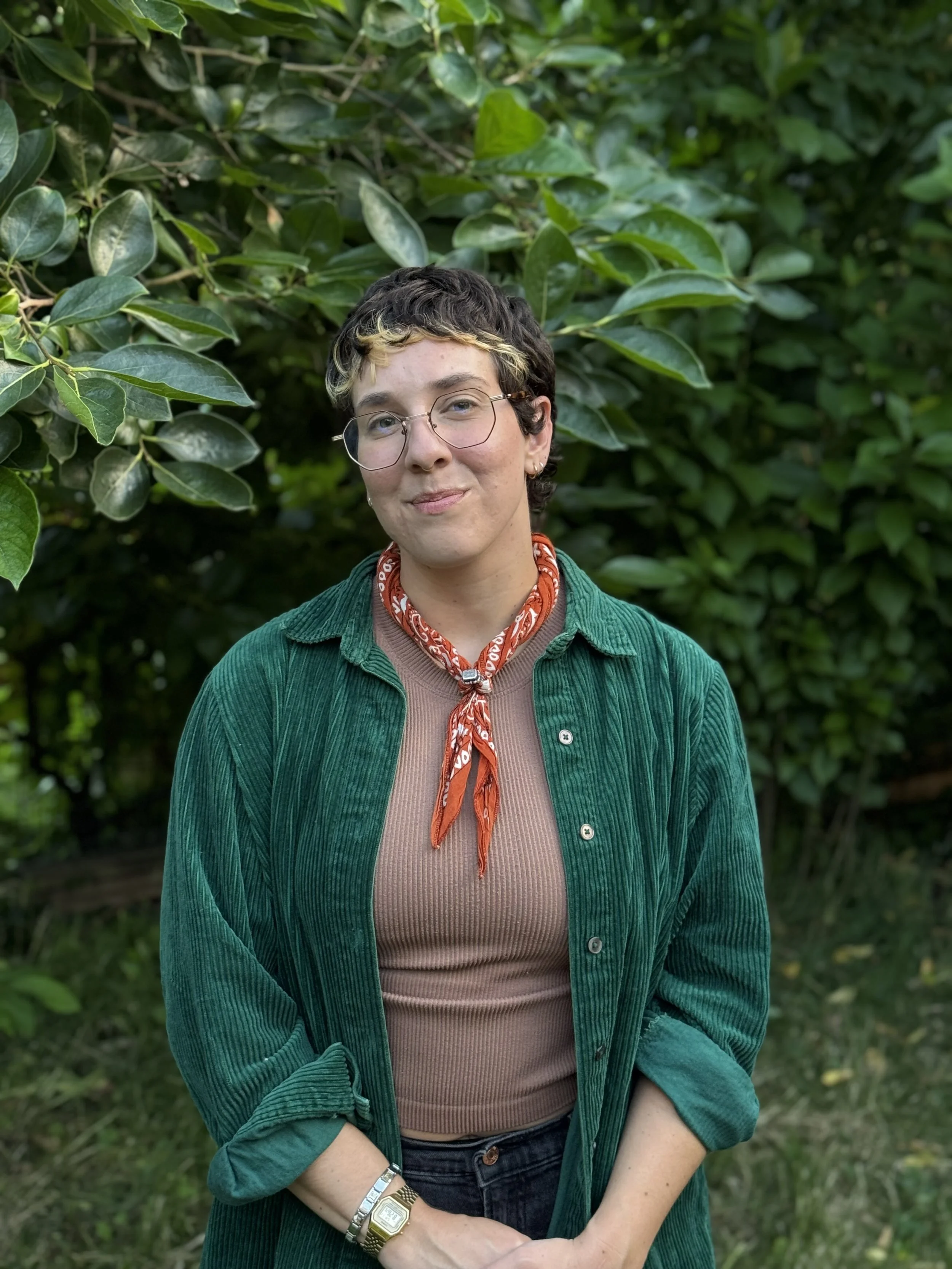 A woman with short, curly dark hair with blonde highlights, wearing glasses, a red bandana, a green corduroy jacket, a brown ribbed shirt, black jeans, and a gold watch, standing outdoors in front of green foliage.