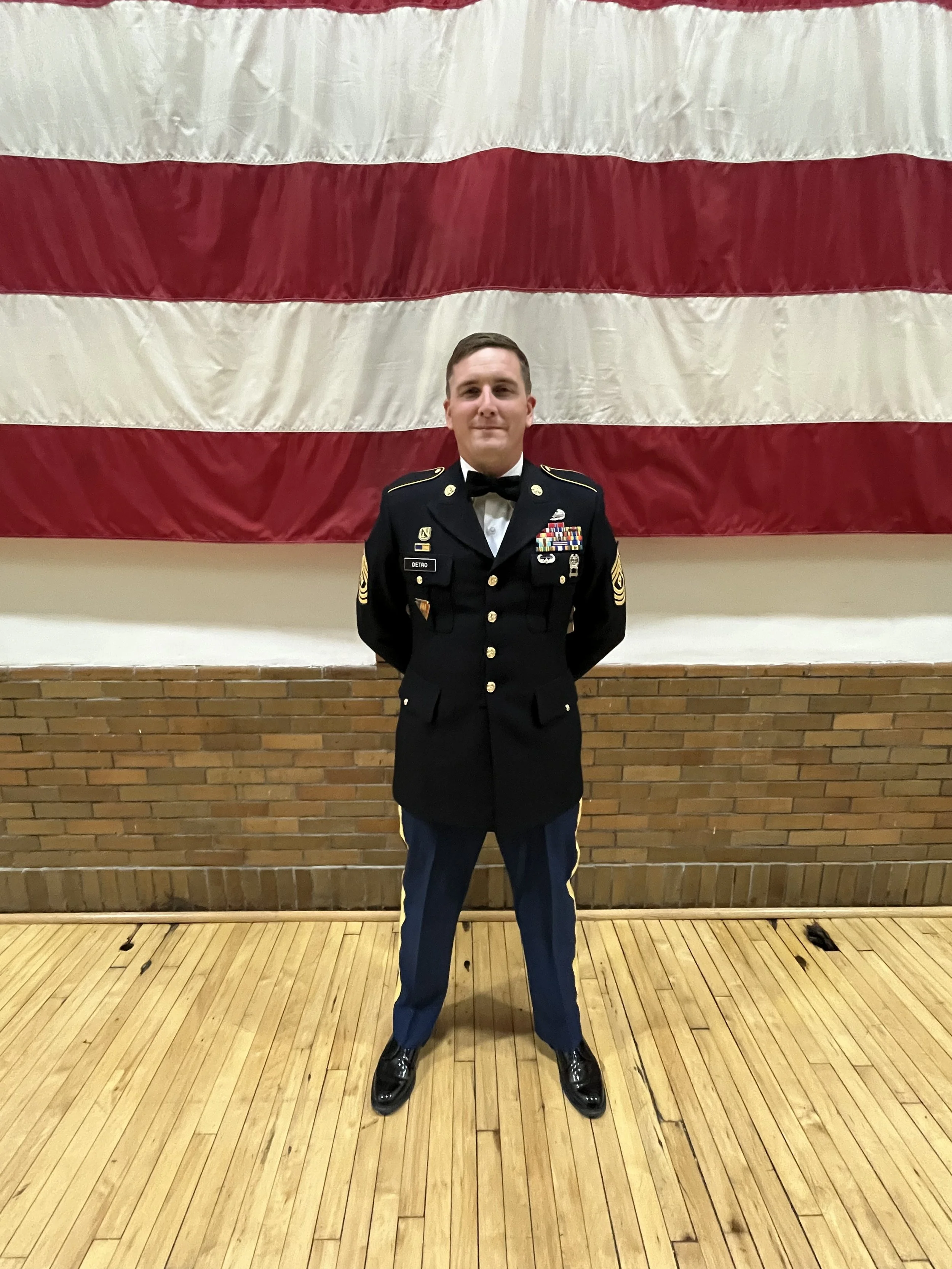 A young man in a dress uniform standing in front of a large American flag, indoors with wooden flooring and a brick wall.