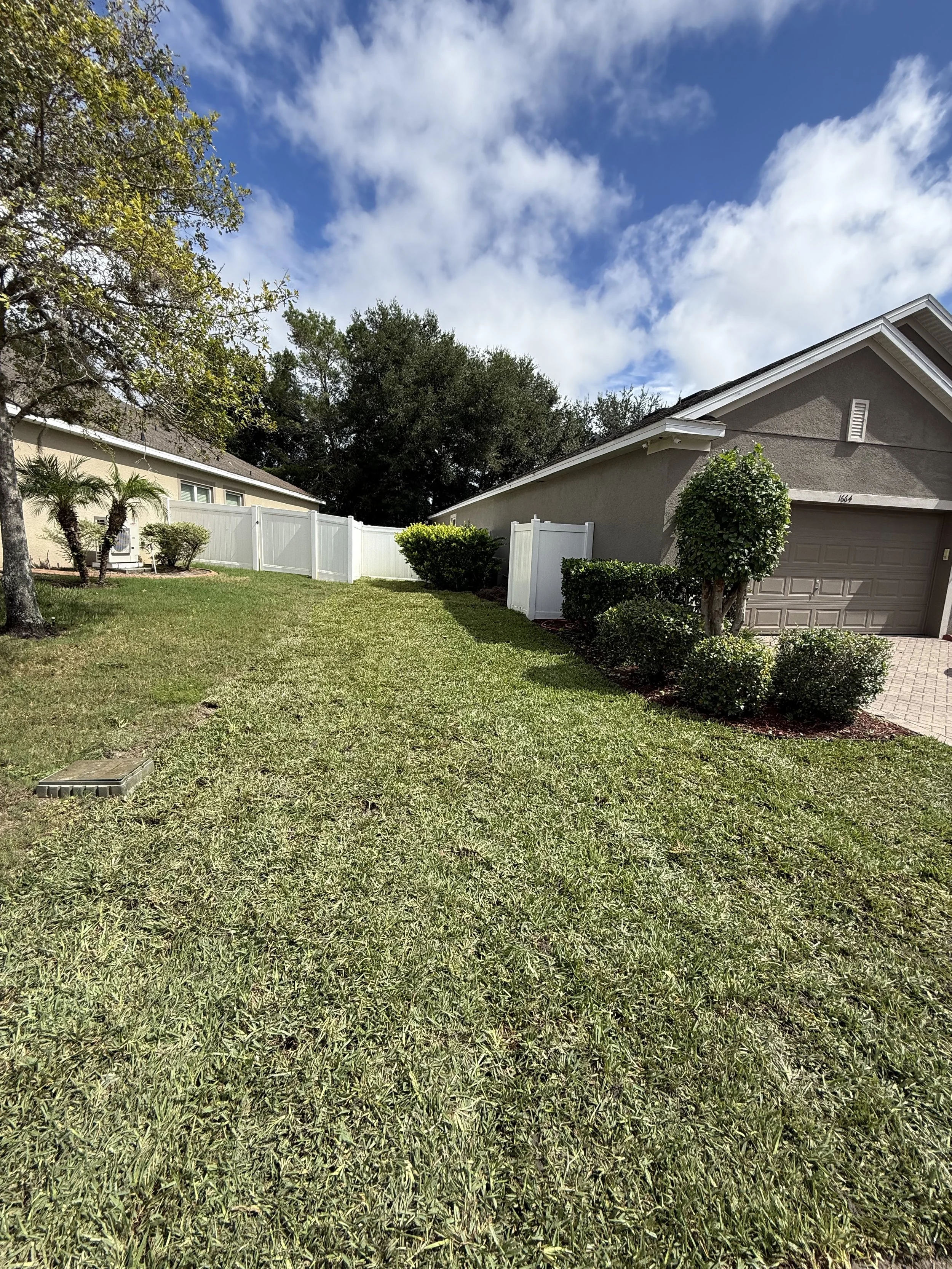 A suburban backyard with trimmed green grass, a large tree on the left, trimmed bushes, a white vinyl fence, and a gray house with a garage door on the right. Blue sky with some clouds is overhead.