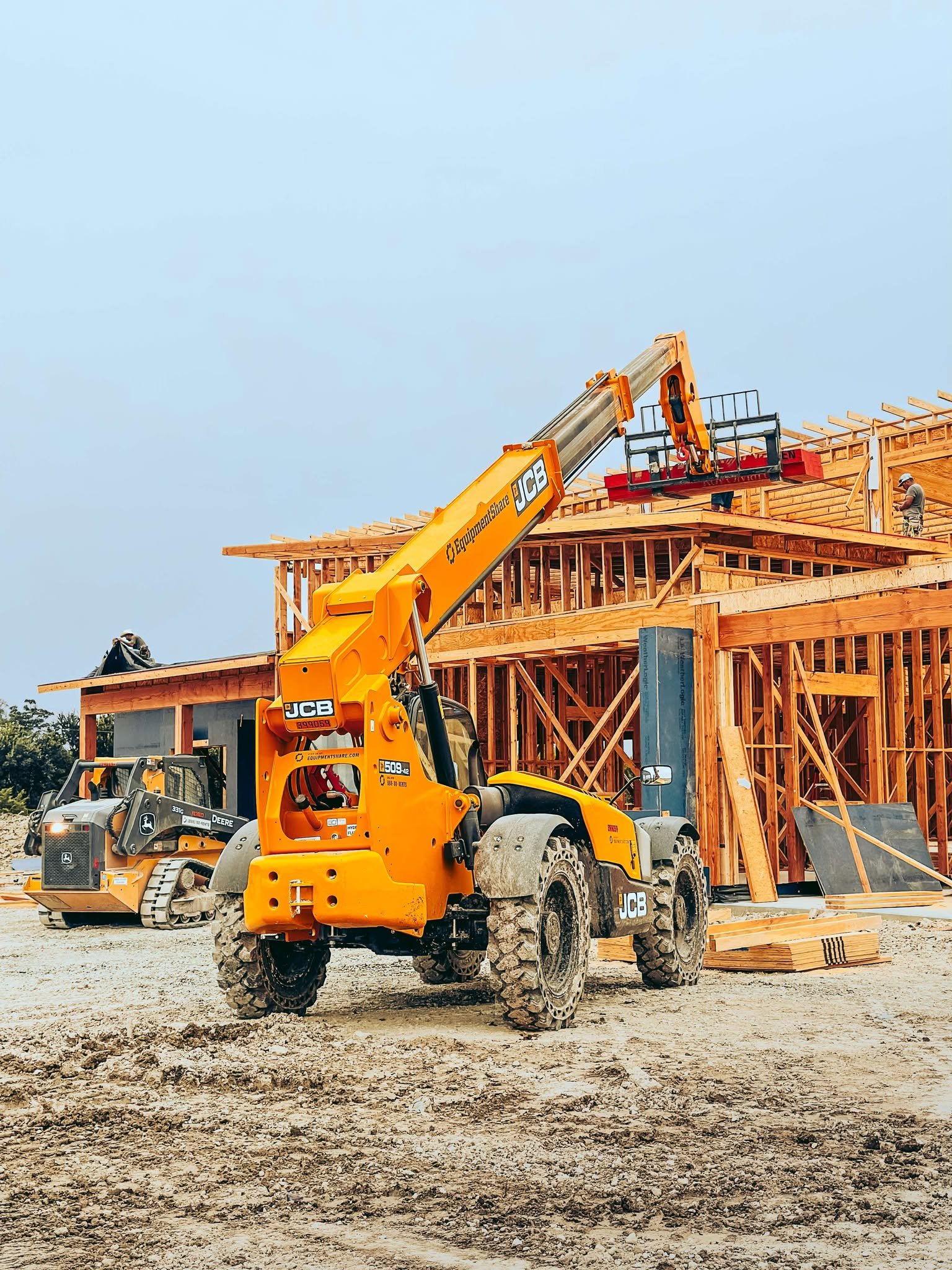 Construction site with a yellow JCB telehandler lifting building materials onto a wooden structure. Another piece of heavy machinery is visible in the background.