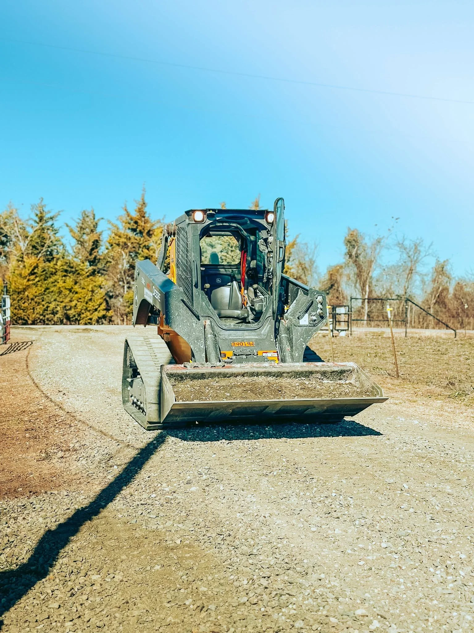 Skid steer loader on a gravel path with trees in the background under a clear blue sky.