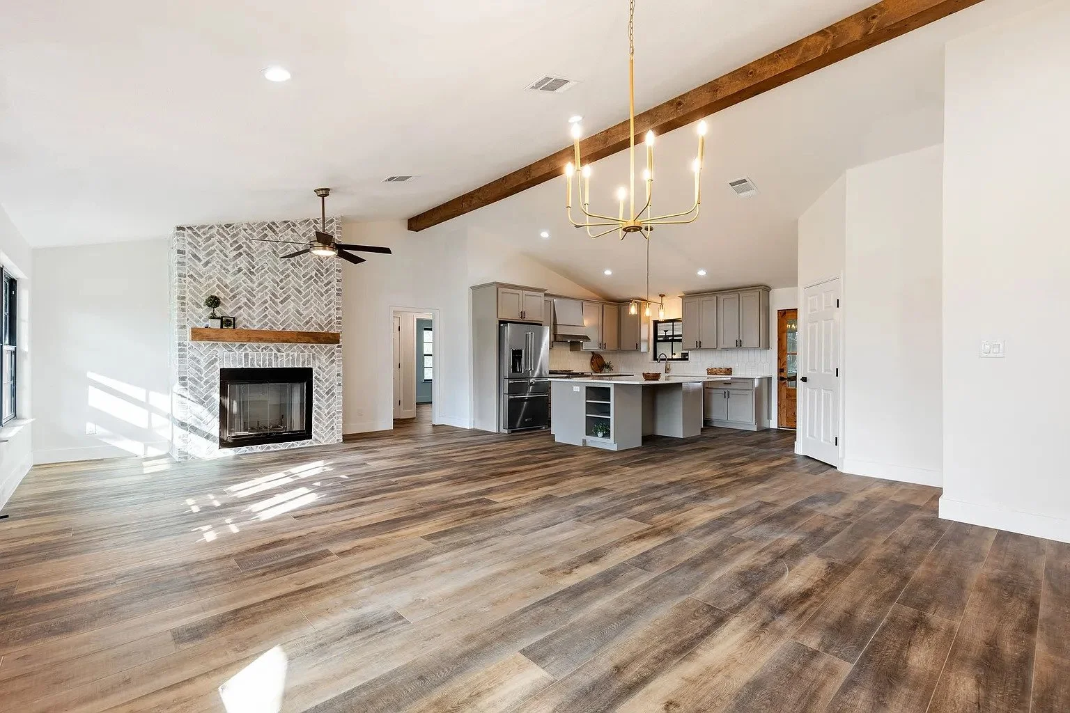 Open concept living area with kitchen and fireplace, featuring wood flooring, vaulted ceiling with beams, and modern lighting fixtures.