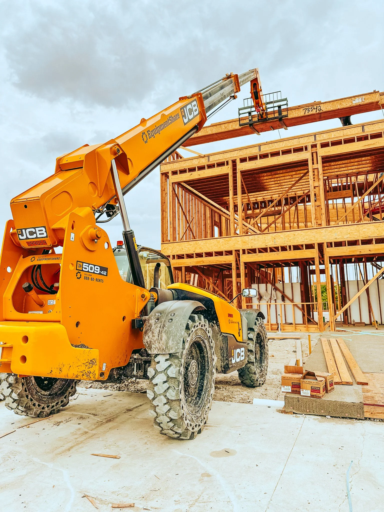 A yellow JCB telescopic forklift at a construction site lifting a wooden beam in front of a wooden-framed building under construction.