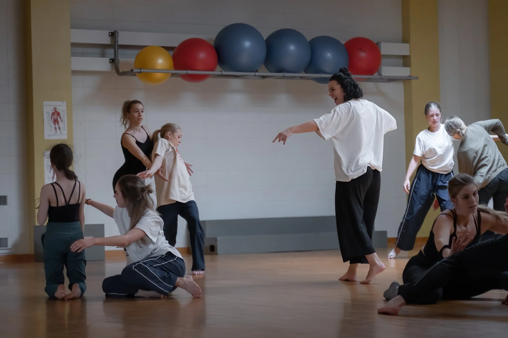 Un groupe de personnes, dont plusieurs jeunes filles, en train de pratiquer la danse ou le yoga dans une salle de sport, sous la supervision d'une femme qui leur donne des instructions.