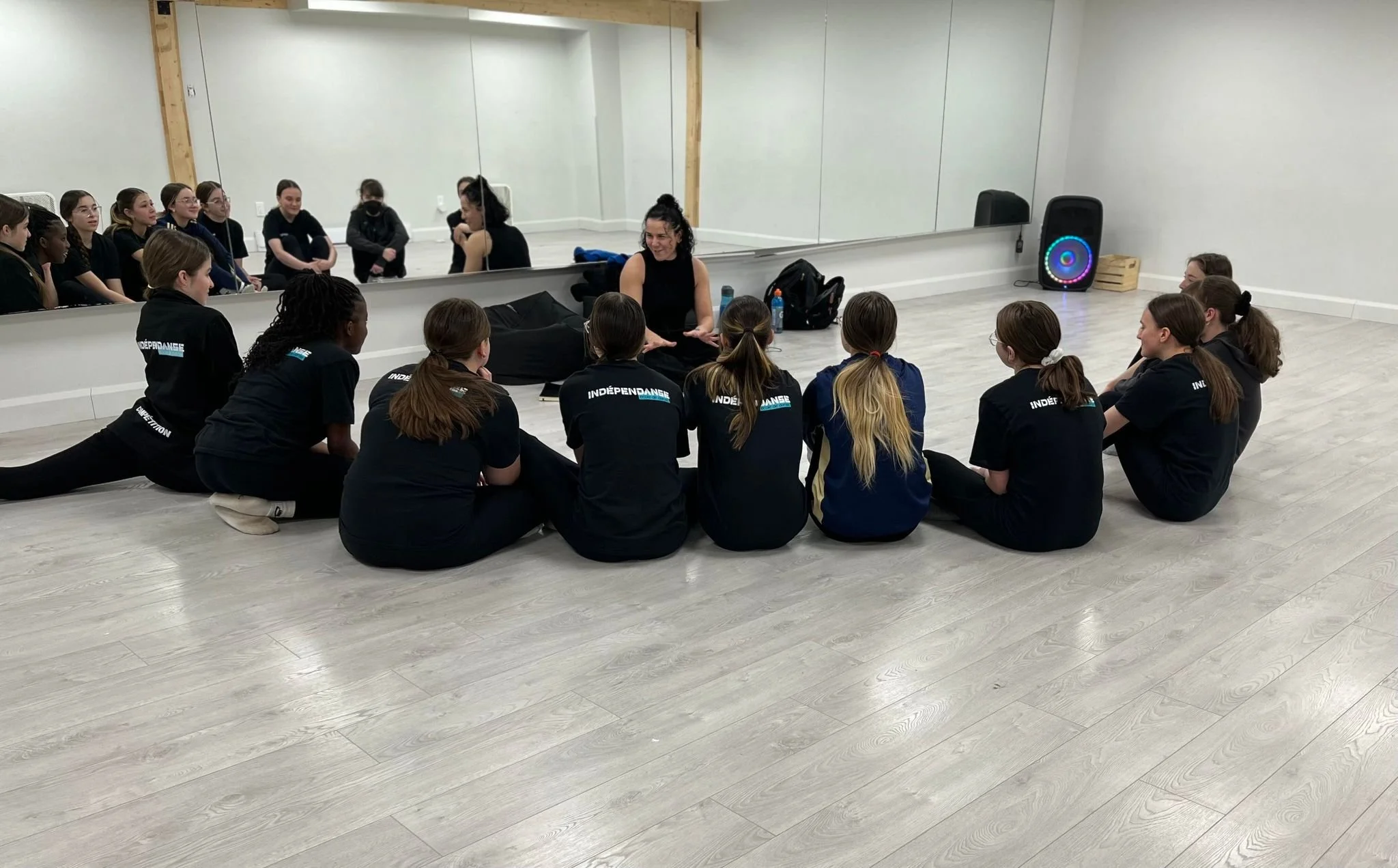 Groupe de jeunes femmes assises en cercle dans une salle de danse, écoutant une femme au centre. Un grand miroir reflète leur image.