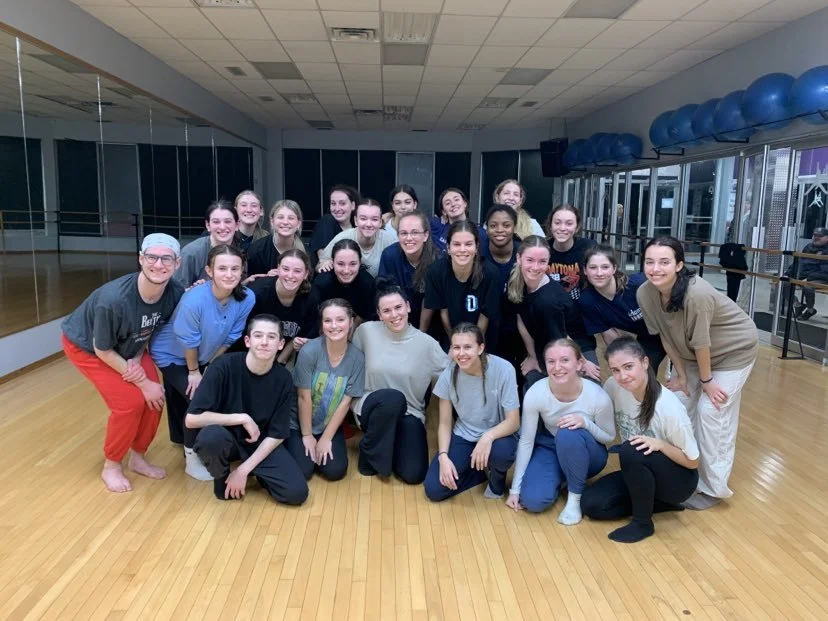 Groupe de jeunes femmes et un homme posant dans une salle de danse ou de sport avec un plancher en bois, miroirs au mur et ballons de fitness en fond.