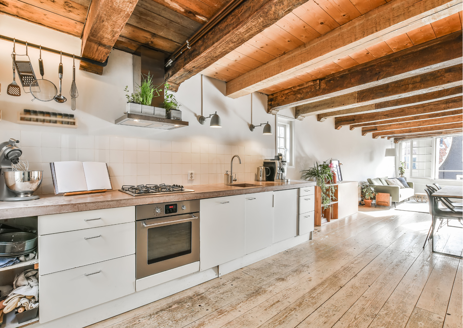A rustic kitchen with wooden beams on the ceiling, white cabinets, stainless steel oven, and a countertop with a faucet. There are hanging utensils and plants, an open book on the counter, and a view of a living area with a sofa and sunlight streamin