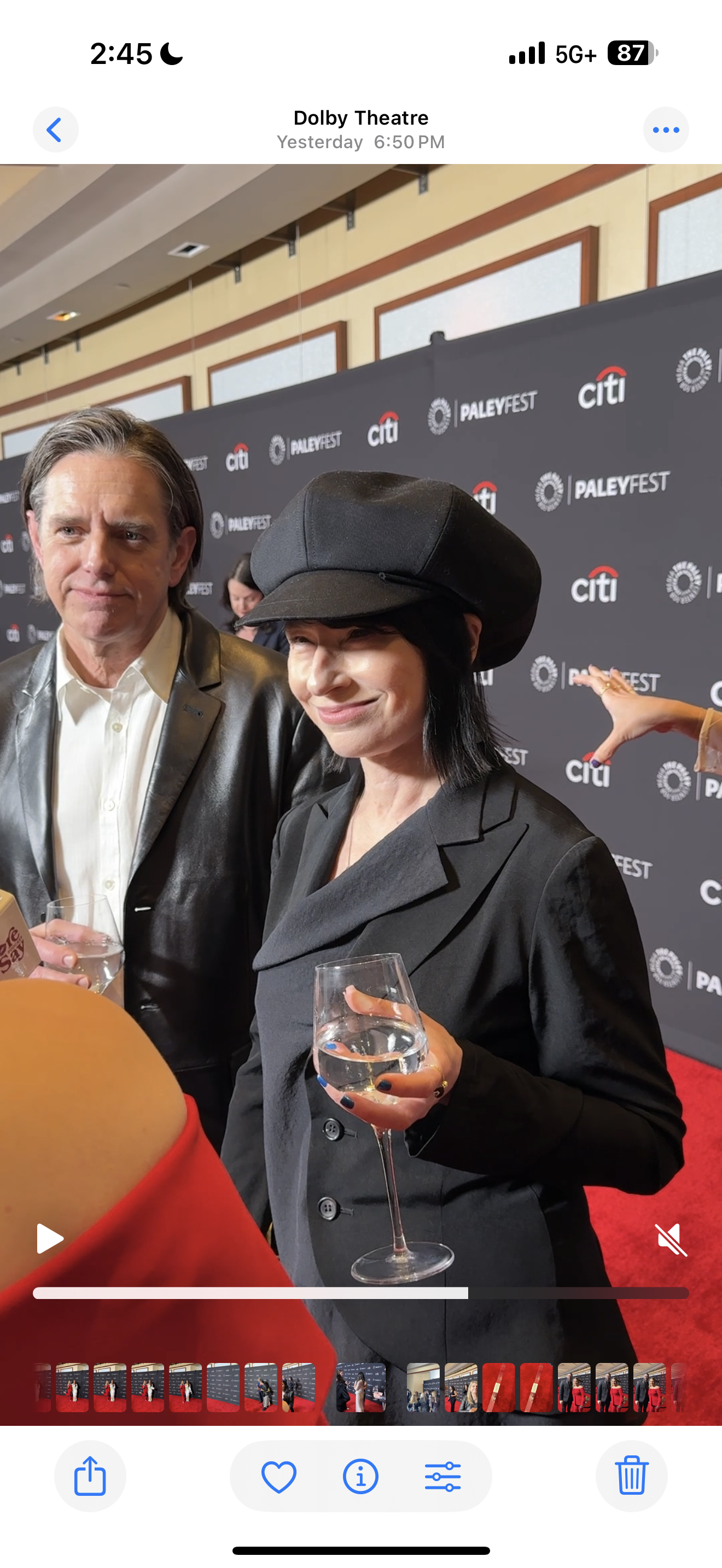 People attending PaleyFest event at Dolby Theatre, with a woman in a black hat and blazer holding a glass of water, and a man in a leather jacket and white shirt beside her, in front of a backdrop with PaleyFest and Citi logos.