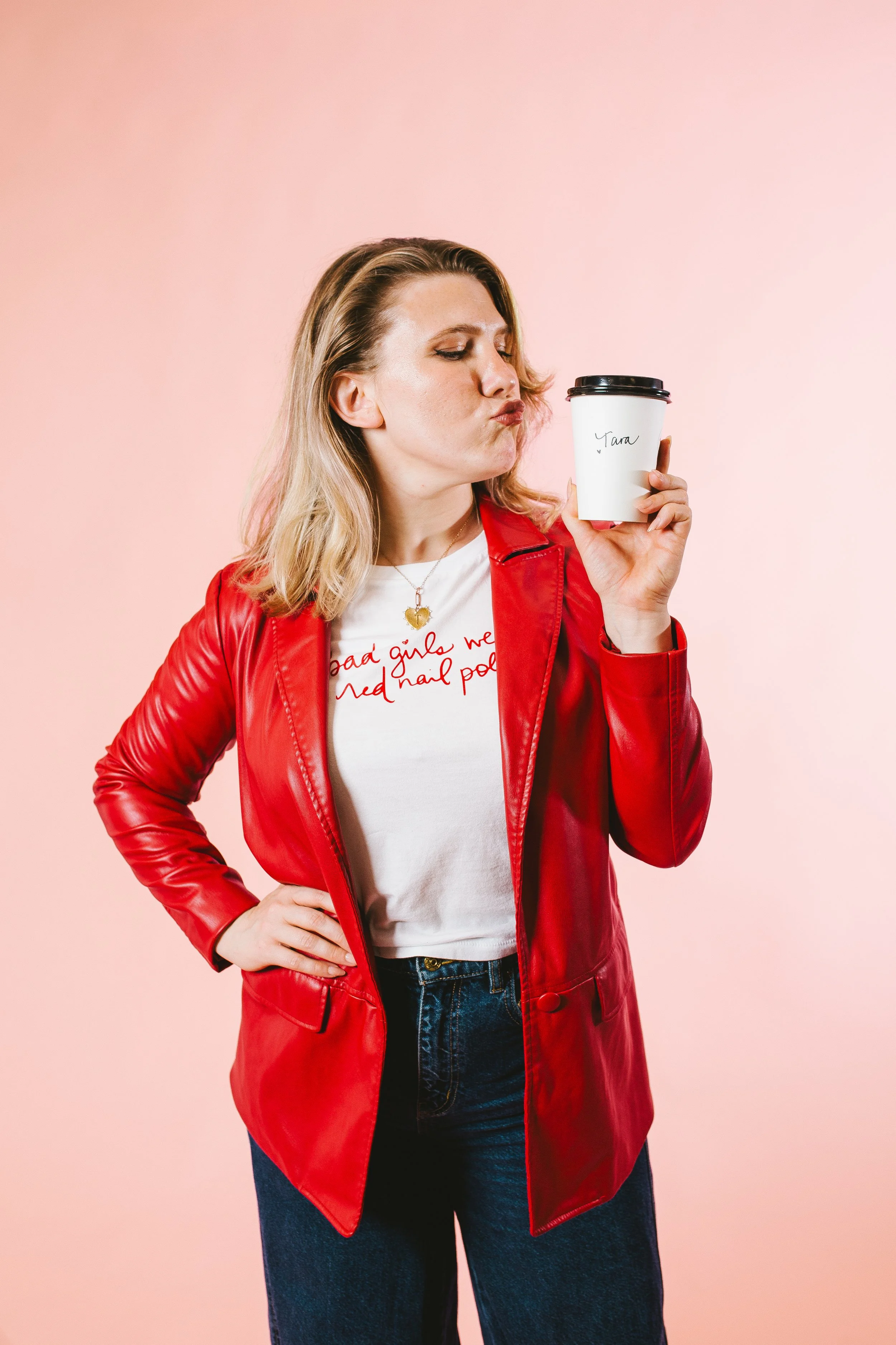 A woman with blonde hair wearing a red leather jacket and white T-shirt holding a coffee cup with her eyes closed and lips puckered against a pink background.