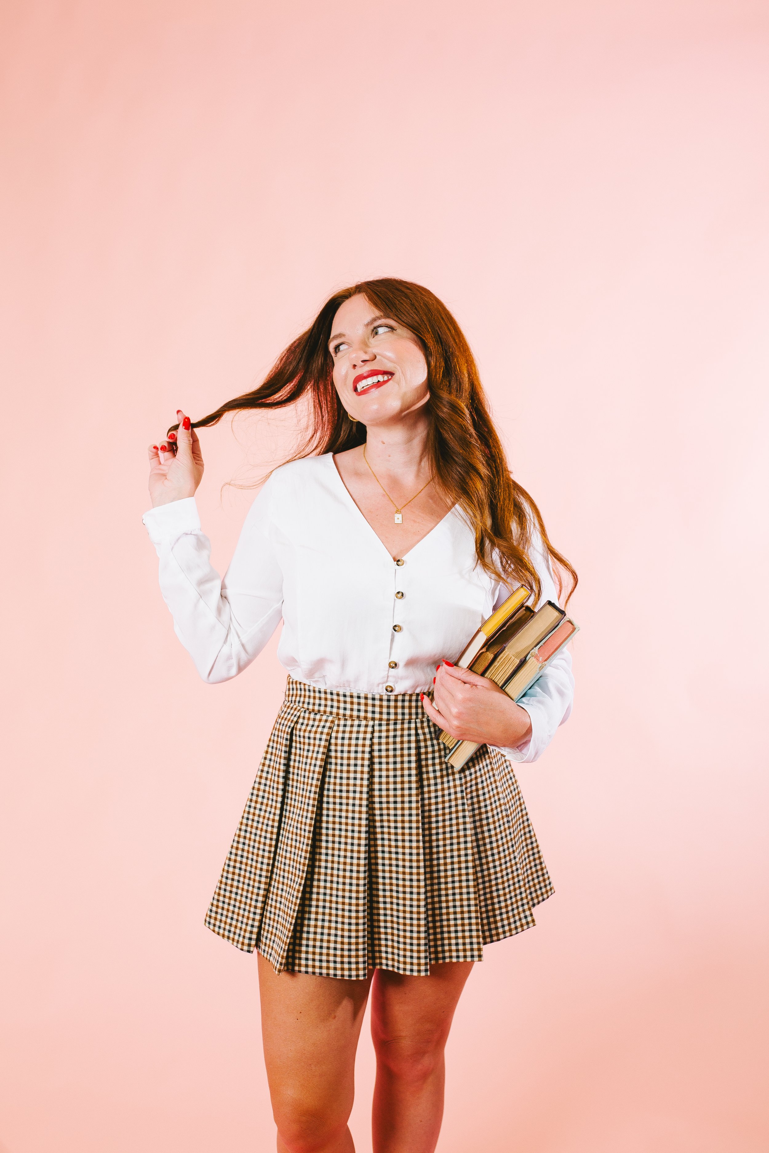 A woman with long, wavy red hair standing against a pink background. She is smiling, holding a few books in one arm, and touching her hair with her other hand. She is wearing a white button-up shirt and a checkered skirt.