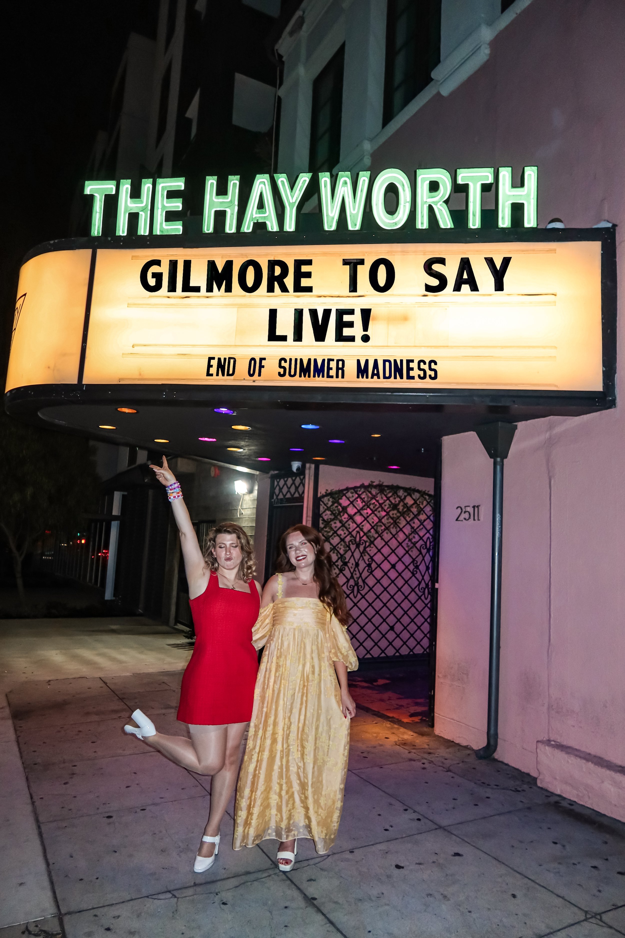 Two women standing in front of a showcase sign outside a theater at night. One woman is wearing a red dress, white heels, and has curly blonde hair, with one arm raised and the other around the other woman. The other woman is wearing a long yellow dress, open-toe heels, and has long auburn hair, smiling at the camera.
