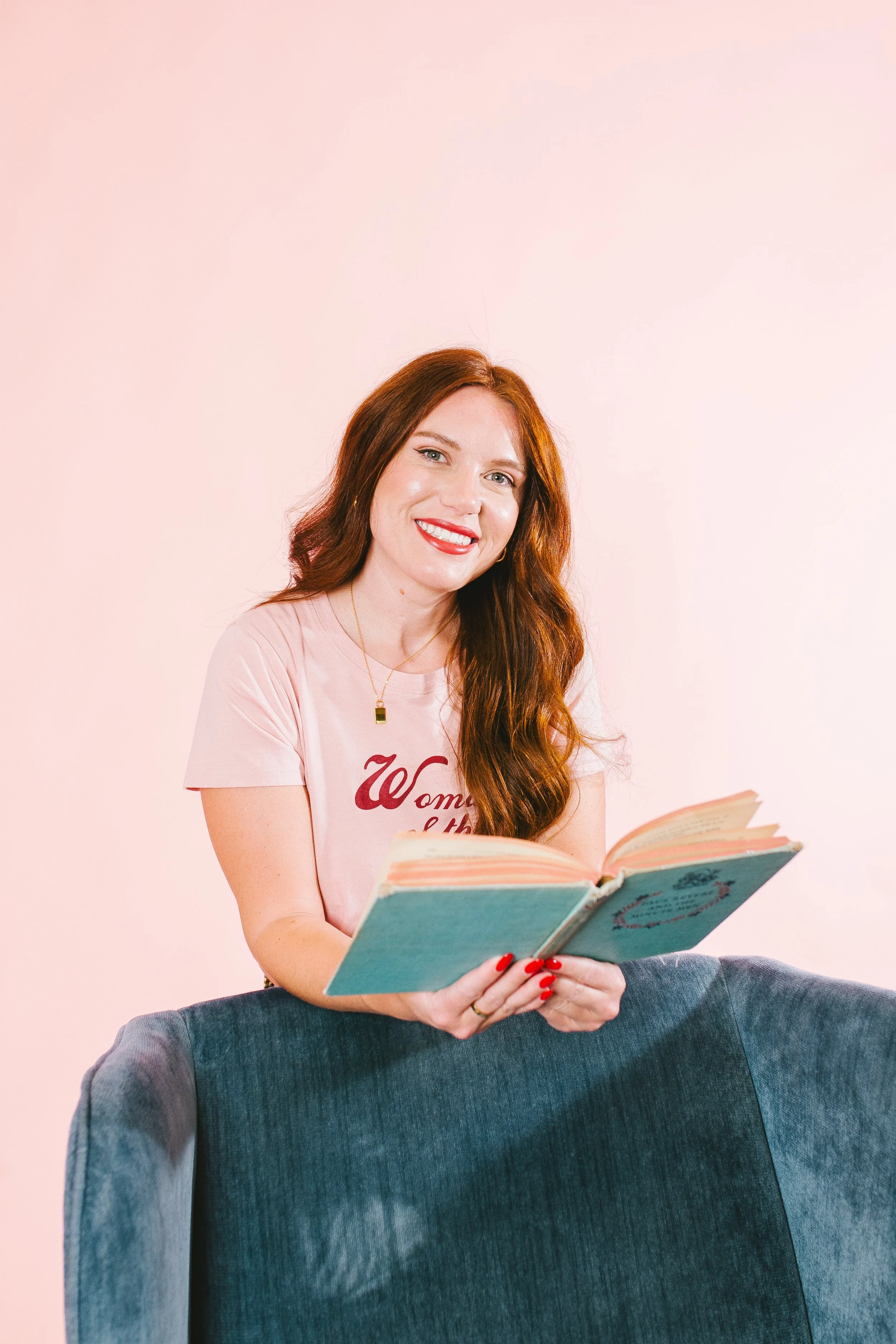 A smiling woman with long red hair, wearing a pink shirt, is sitting on a blue upholstered chair and holding an open book.