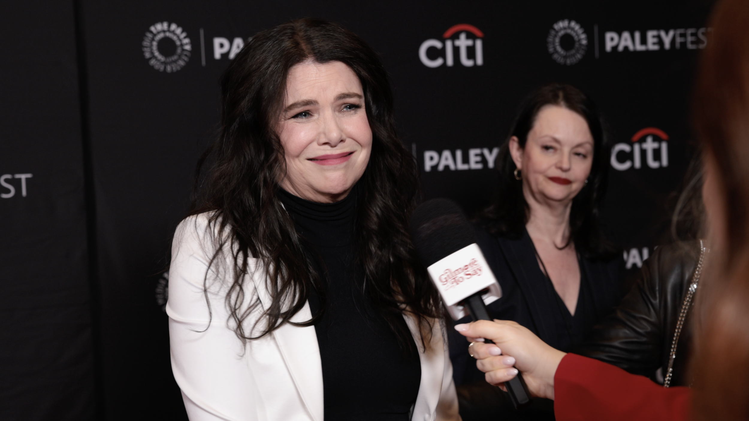 A woman with long dark hair, wearing a black turtleneck and white blazer, is being interviewed with a microphone that says 'Galaxy Bold'. In the background, there is a black backdrop with logos, including 'Paley' and 'Citi'. Another woman with dark hair and red lipstick is standing behind her, looking to the side.