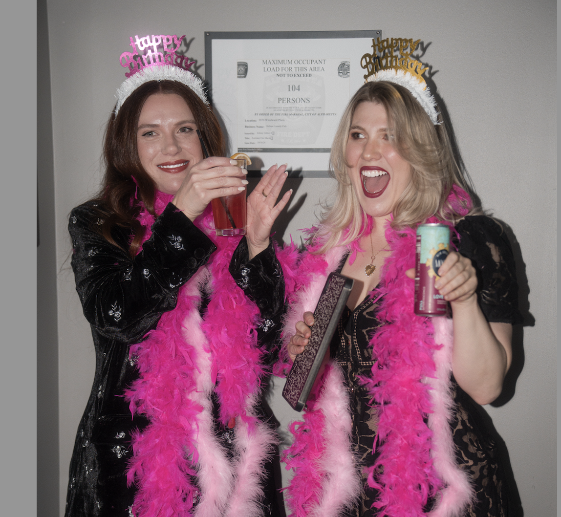 Two women at a birthday party wearing 'happy birthday' tiaras and pink feather boas, smiling and holding drinks, standing in front of a fire department sign.