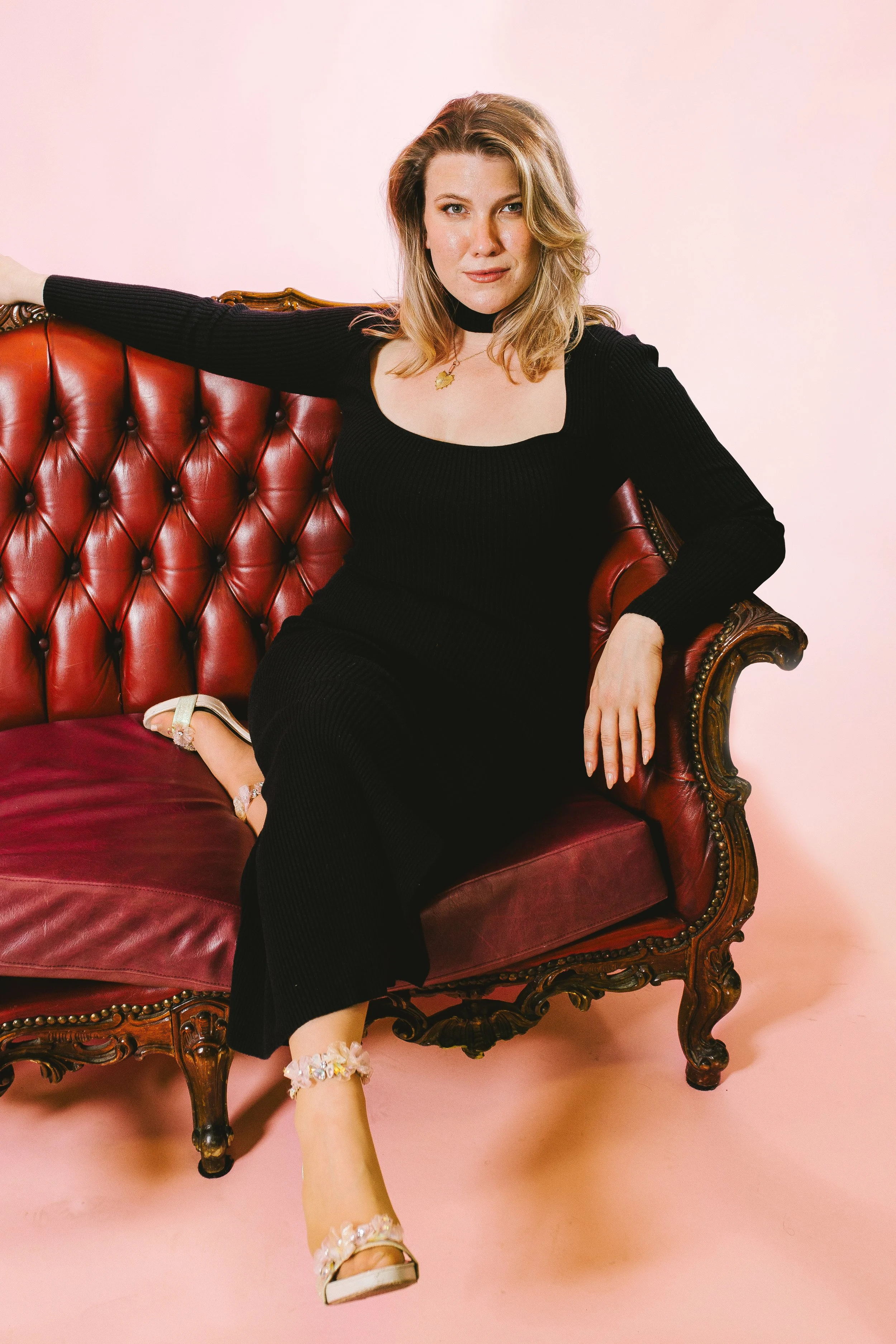 Young woman in a black dress sitting on a vintage red velvet sofa with ornate wooden details, against a pale pink background.