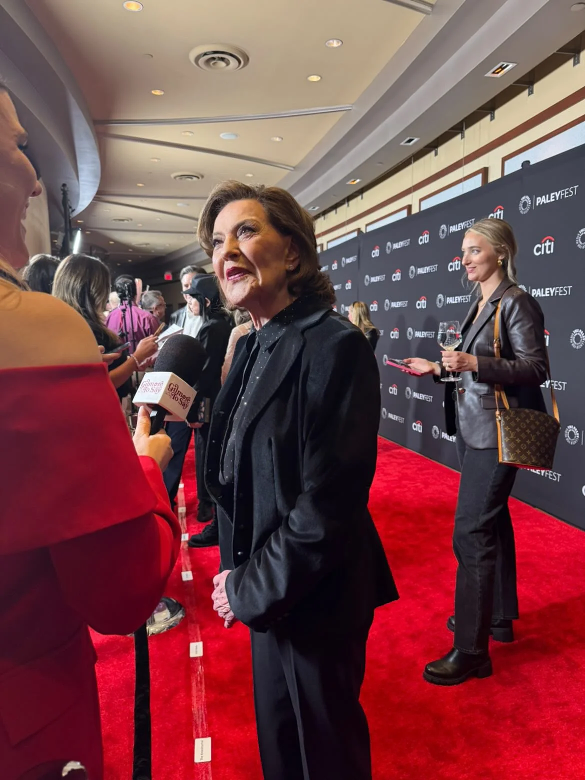 A woman being interviewed with a microphone at an event with a red carpet and a backdrop that reads 'PaleyFest' and 'Citi' behind her. Another woman stands in the background holding a glass of wine.