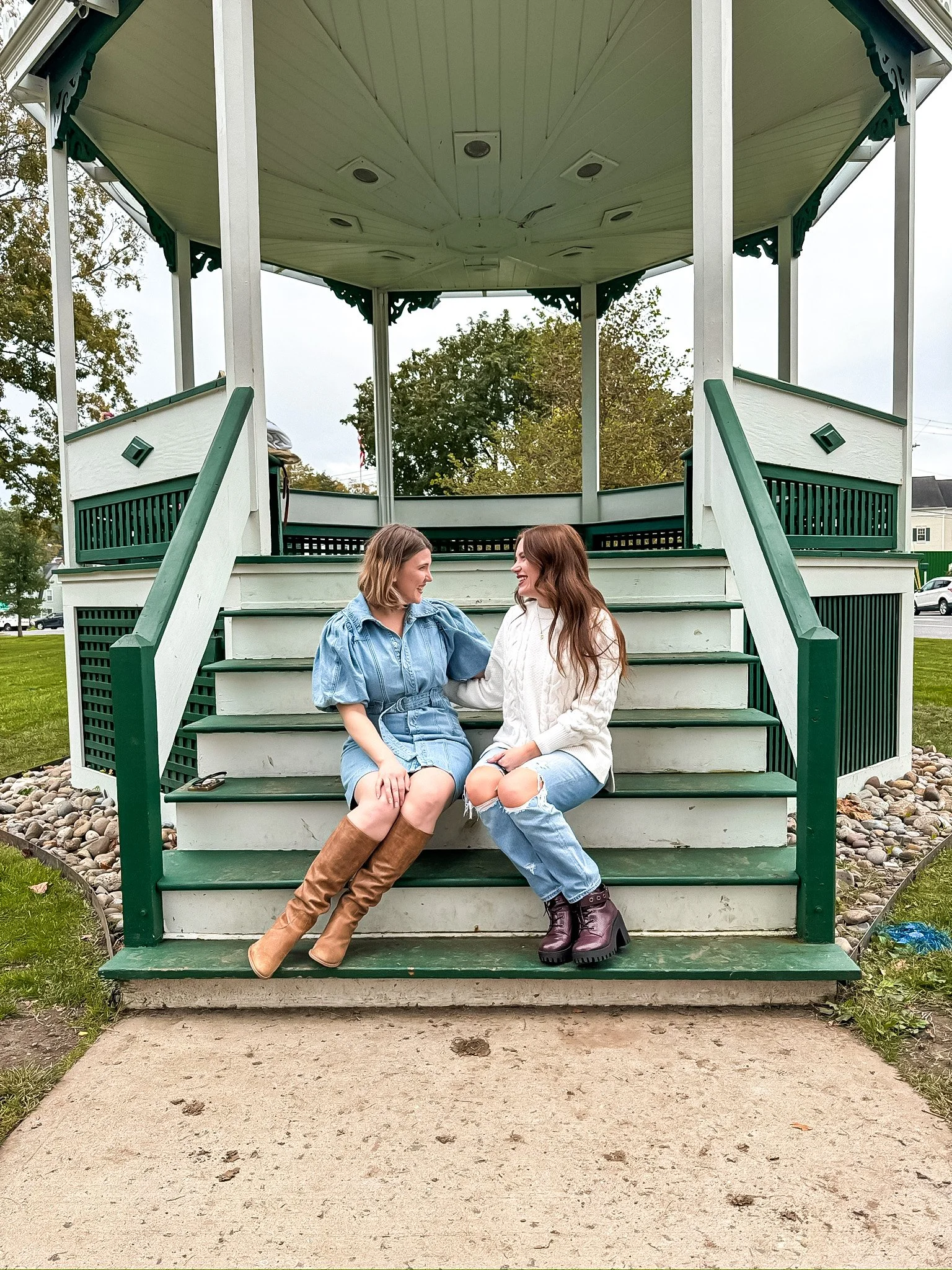 Two women sitting on the steps of a green and white gazebo, smiling and talking to each other, outdoors on a cloudy day.