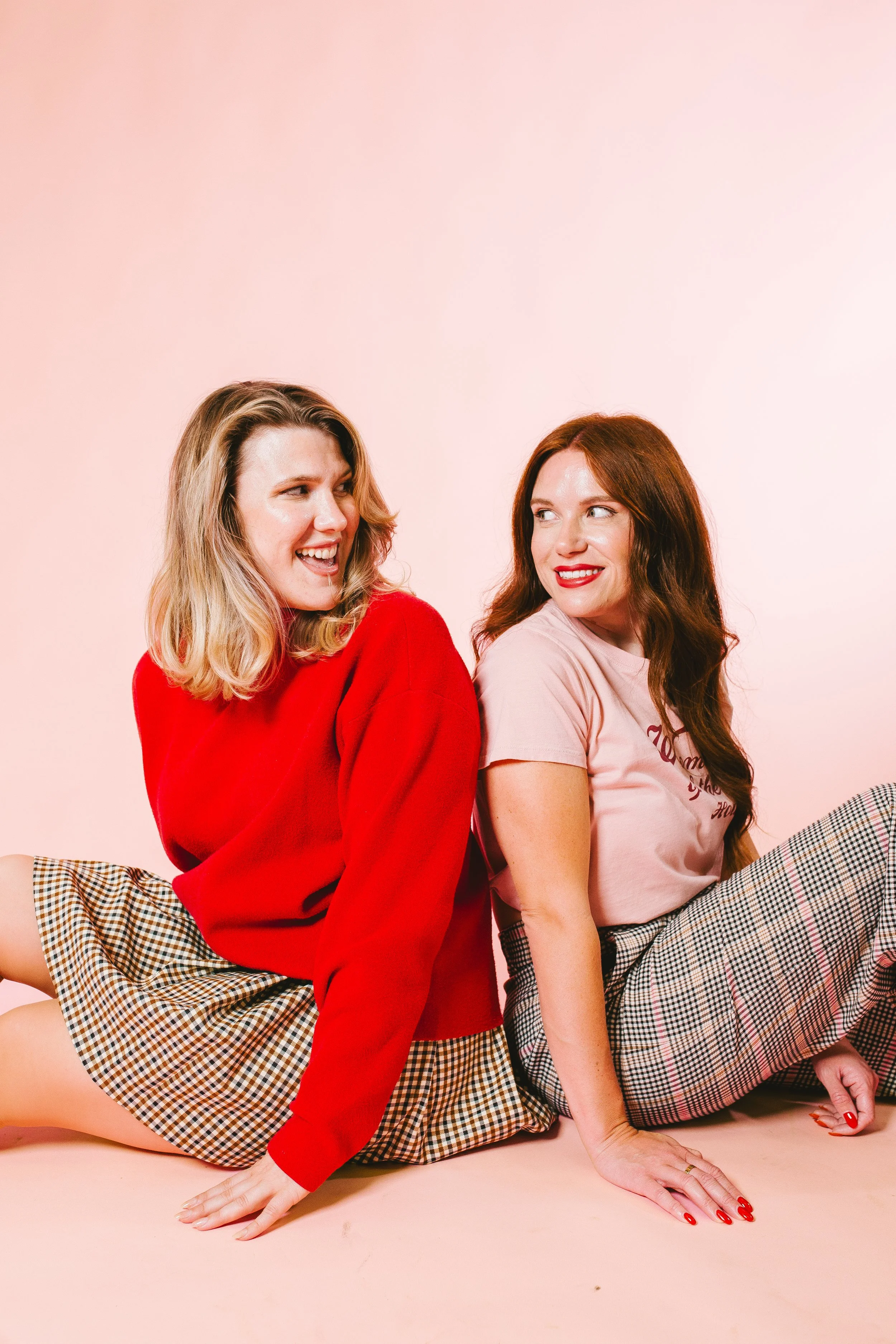 Two women sitting on the floor back-to-back, smiling and laughing, against a light pink background.