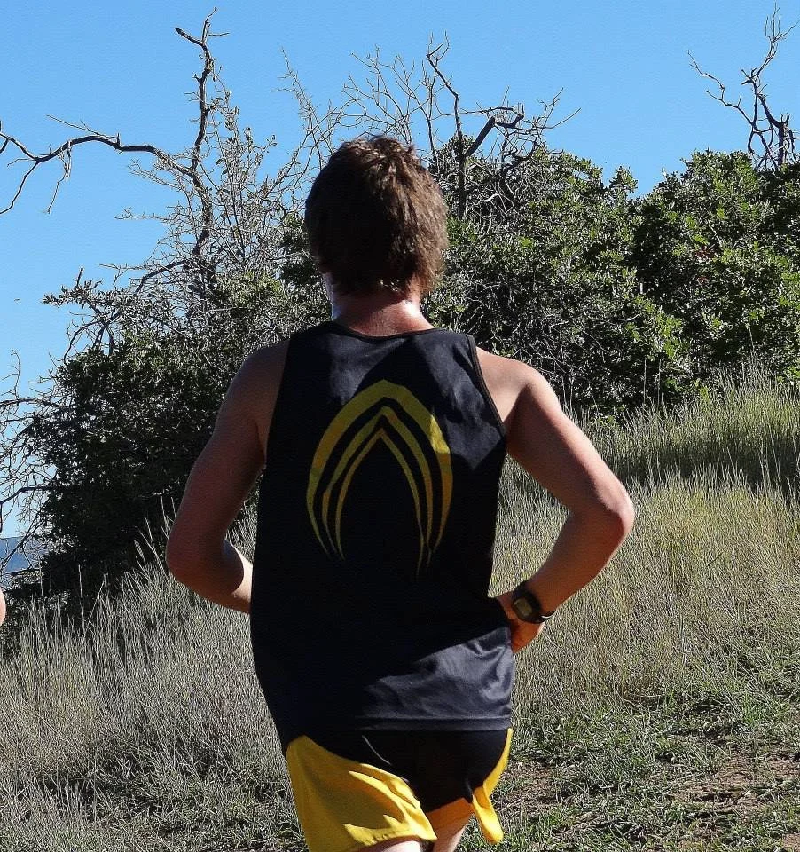 A person with short hair wearing a black sleeveless shirt with yellow design on the back and yellow shorts, running on a trail through a grassy area with trees and leafless branches under a clear blue sky.