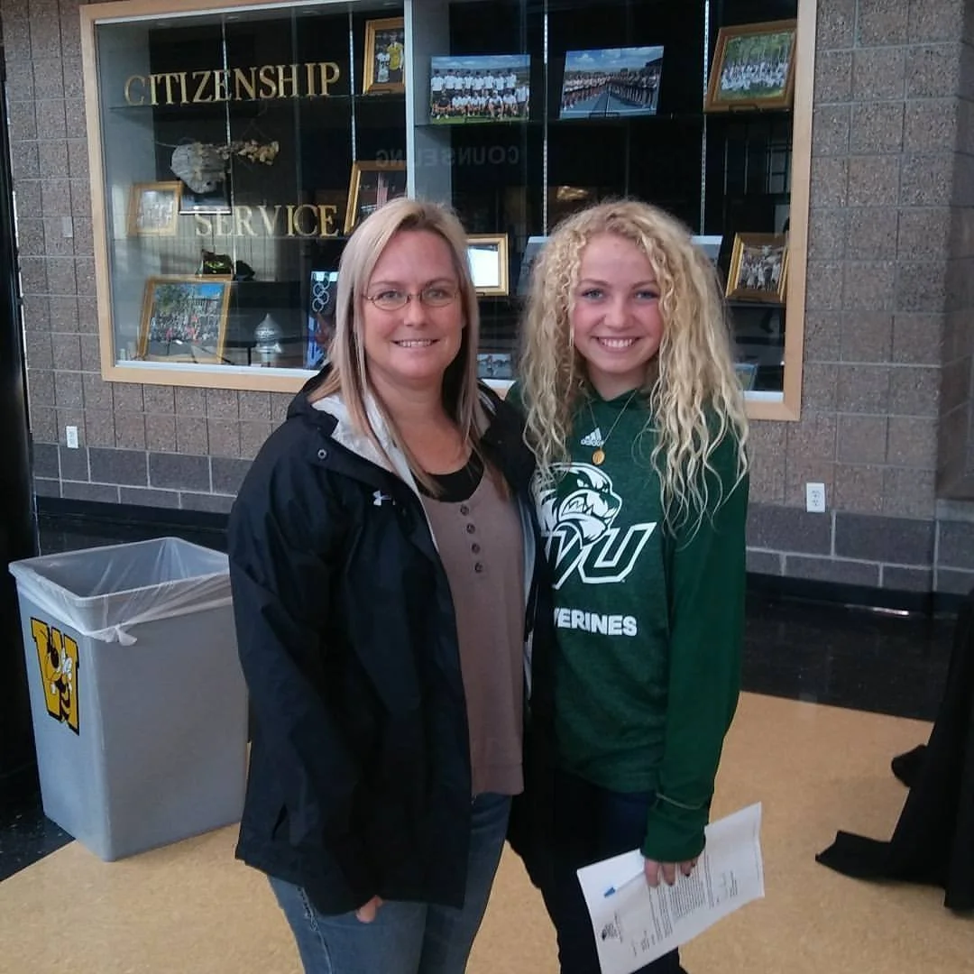 Two women standing together indoors, smiling at the camera. The woman on the right is wearing a green athletic shirt with a lion logo and the word "Warriors." The background includes a display case with photos and the words "CITIZENSHIP" and "SERVICE" visible.