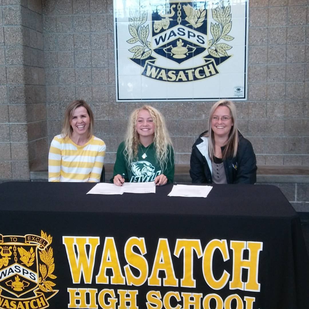 Three women sitting at a table with a "WASATCH HIGH SCHOOL" banner, two have papers in front of them, behind a wall with a large Wasatch logo sign.