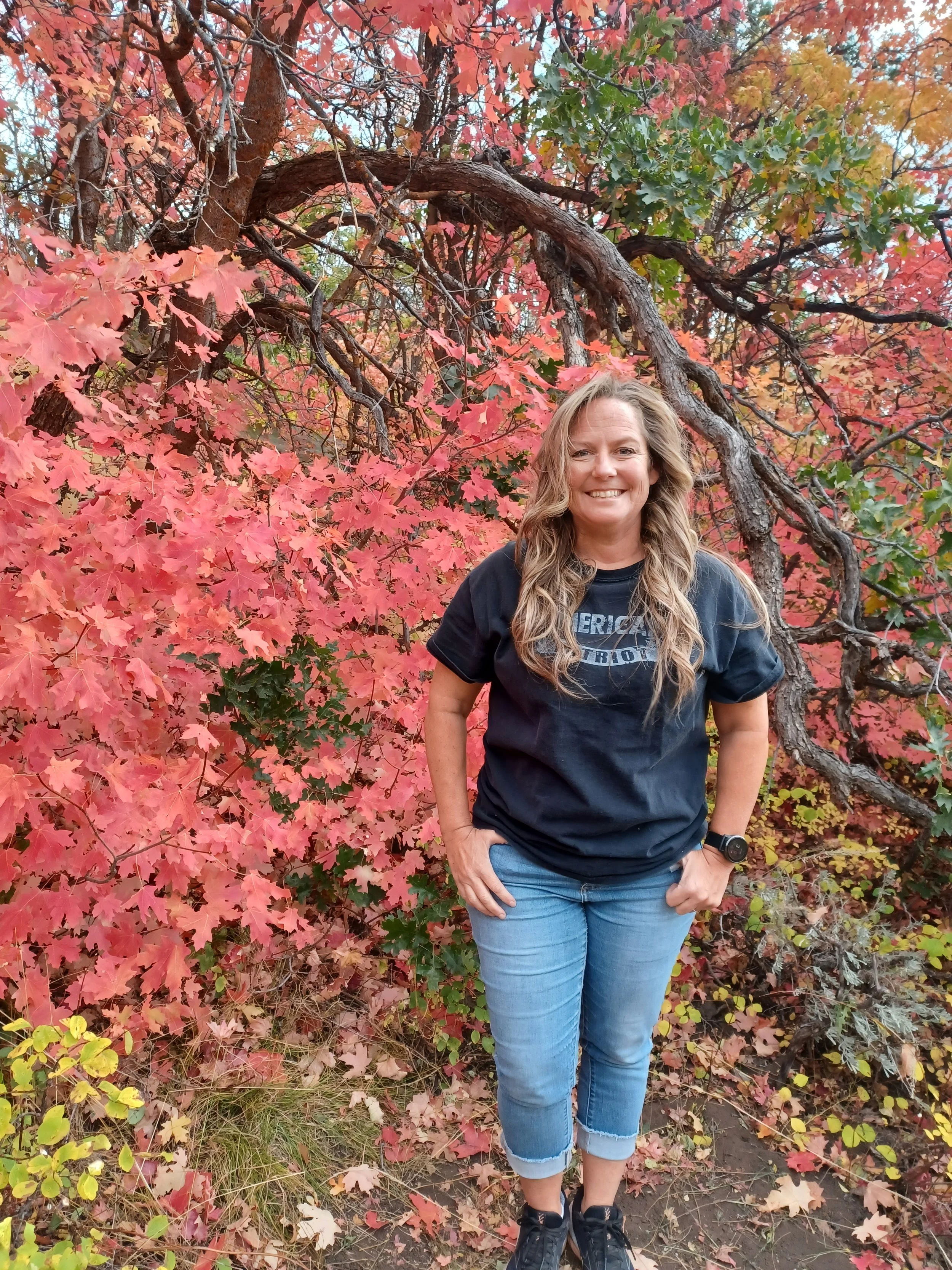 A woman standing outdoors surrounded by colorful autumn leaves in shades of pink, red, and orange.