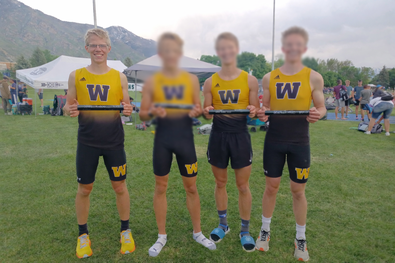 Four male track athletes in yellow and black uniforms hold batons, standing on a grassy field with tents and people in the background, mountains visible in the distance.