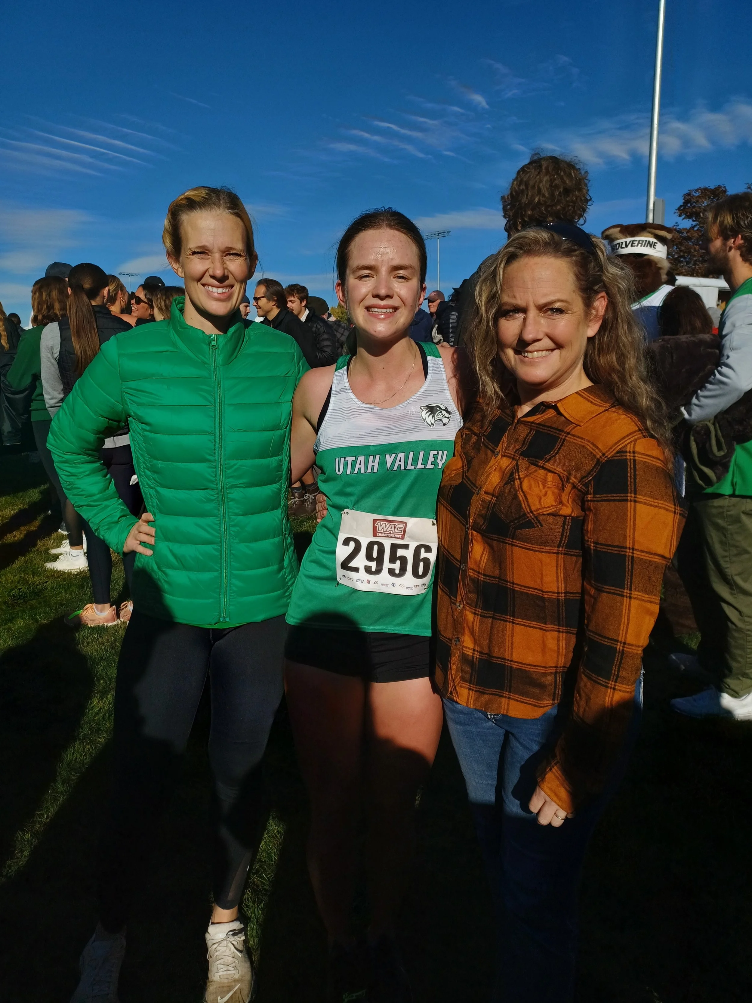 Three women smiling at a race event, with a crowd in the background and a blue sky overhead.