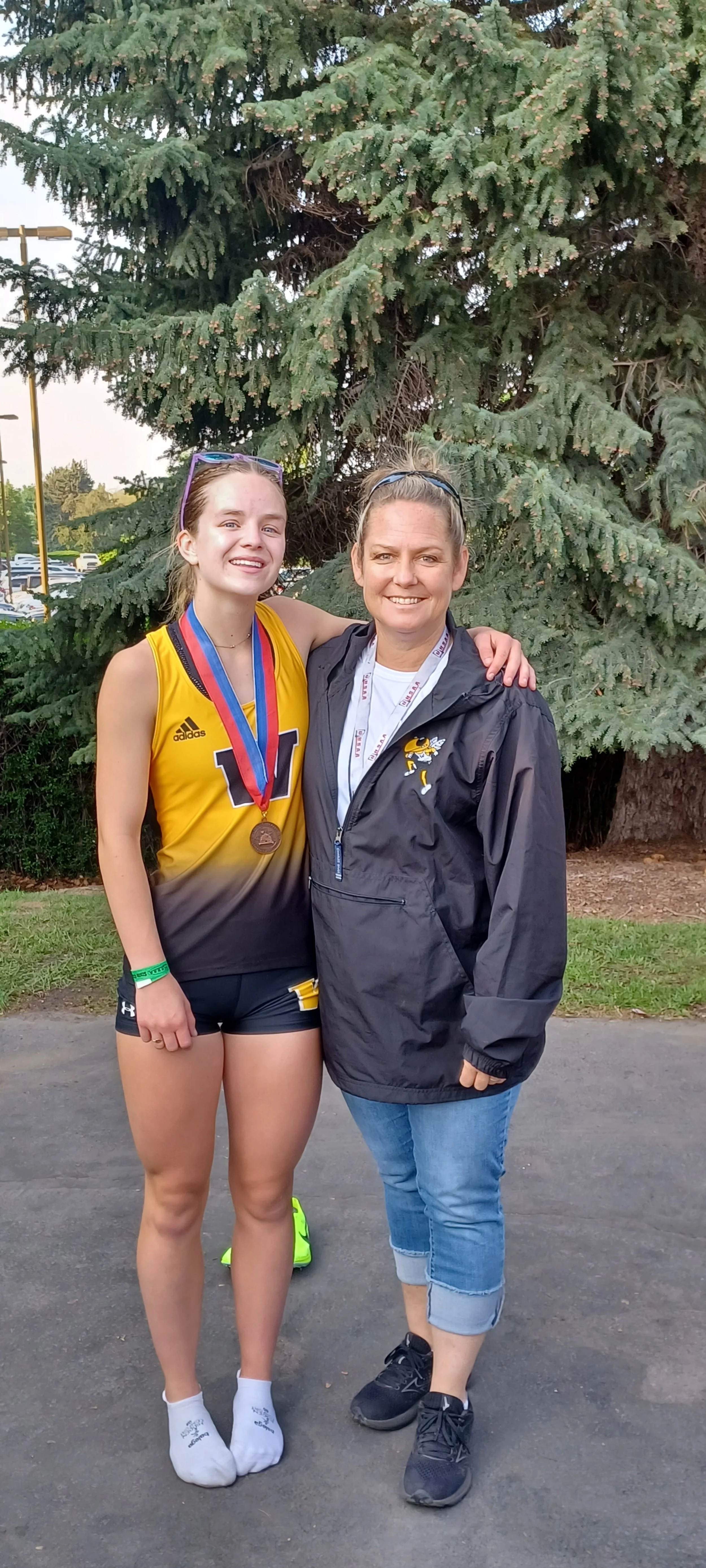 A young woman wearing a yellow and black athletic uniform with medals around her neck and an older woman in a black jacket and jeans posing together outdoors in front of a large evergreen tree.