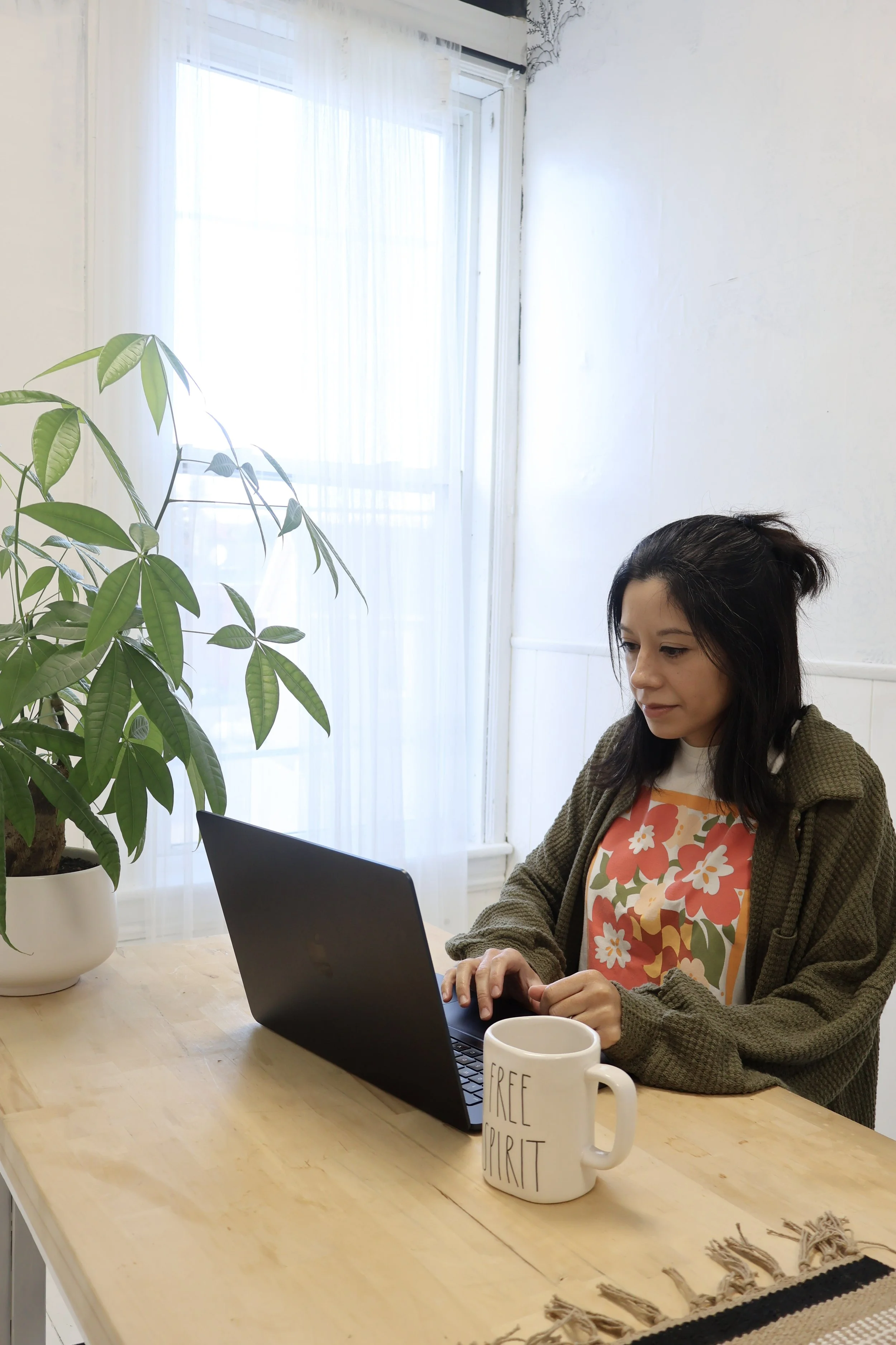 A young woman with dark hair working on a laptop at a wooden table in a bright room with white walls and a large window with sheer curtains. A green potted plant is on the table and a white mug with the phrase 'Free Spirit' printed on it is also on the table.