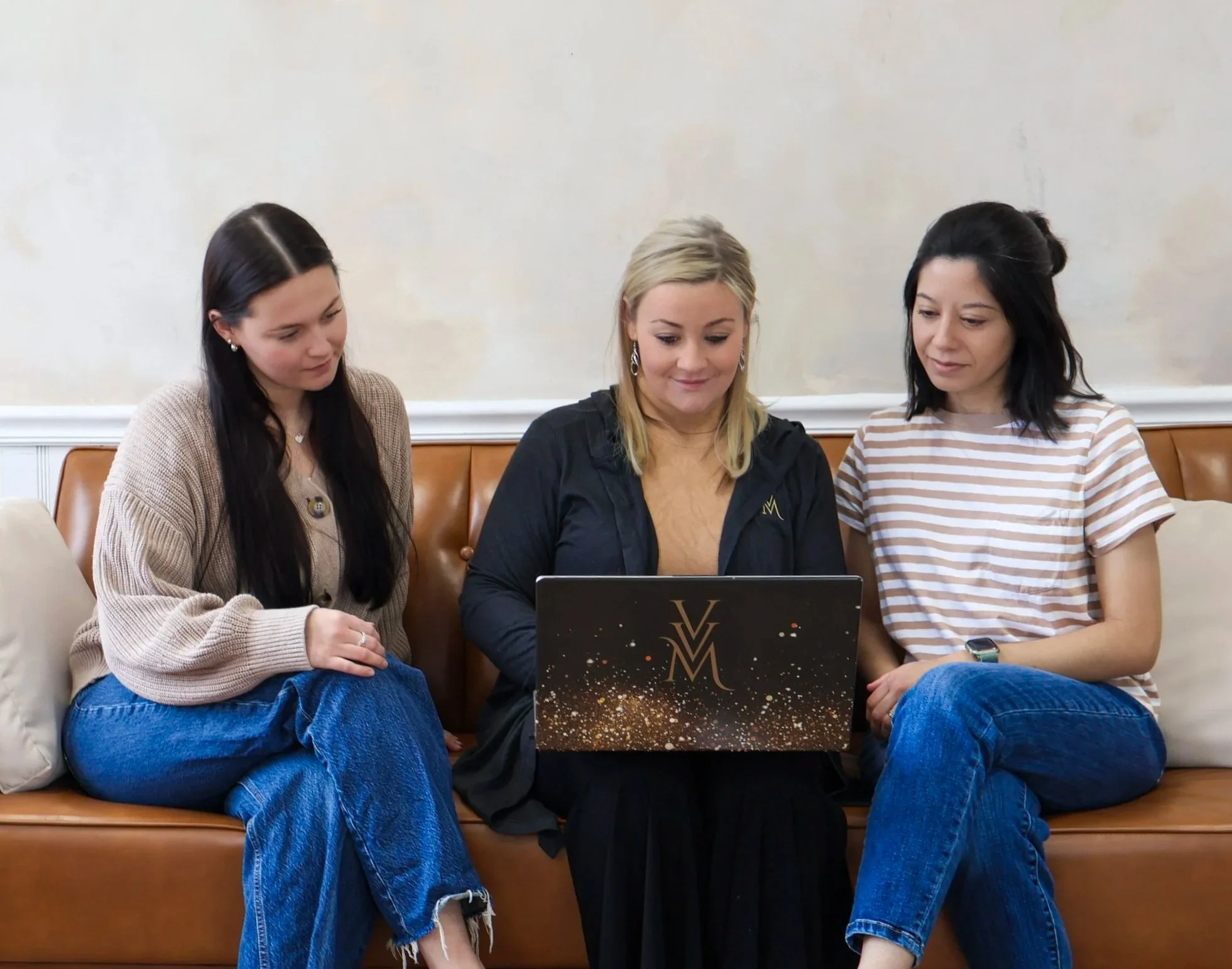 Three women sitting closely together on a brown leather couch, looking at a laptop with a model Y logo, in a room with light-colored walls.