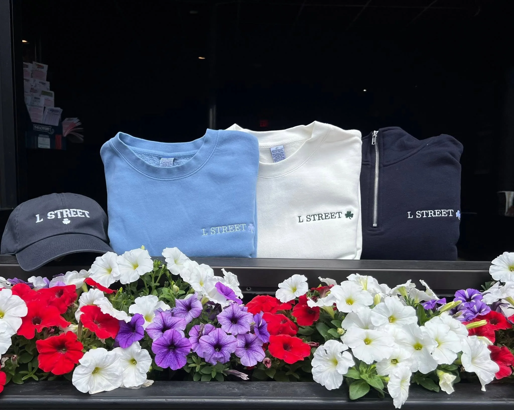 Display of three folded T-shirts in light blue, white, and black, each embroidered with 'L STREET' and a small shamrock, arranged above a bed of colorful petunias.
