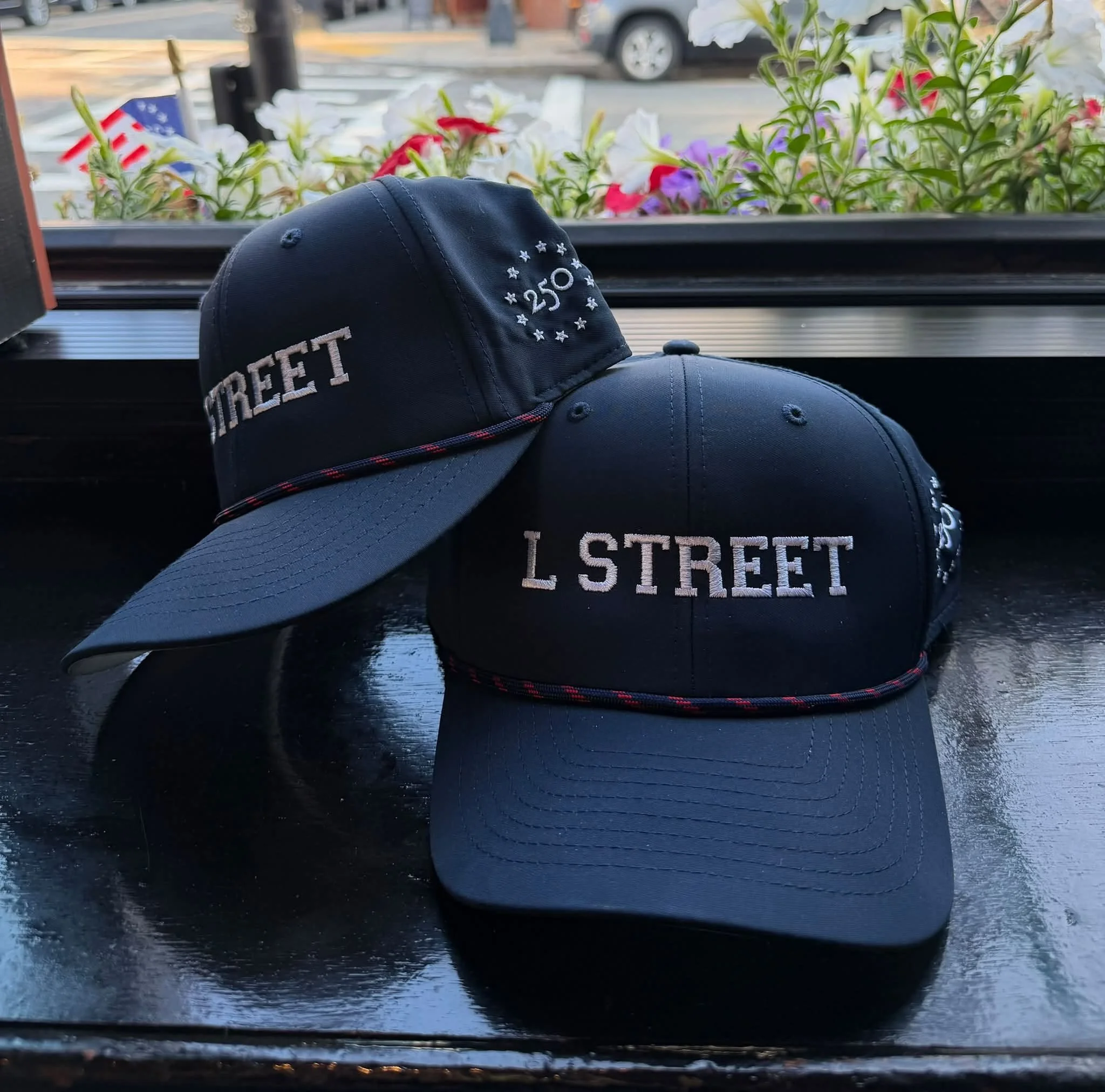 Close-up of a navy blue baseball cap with the text 'L STREET' and the number '250' embroidered on it, resting against a window with a flower planter outside.