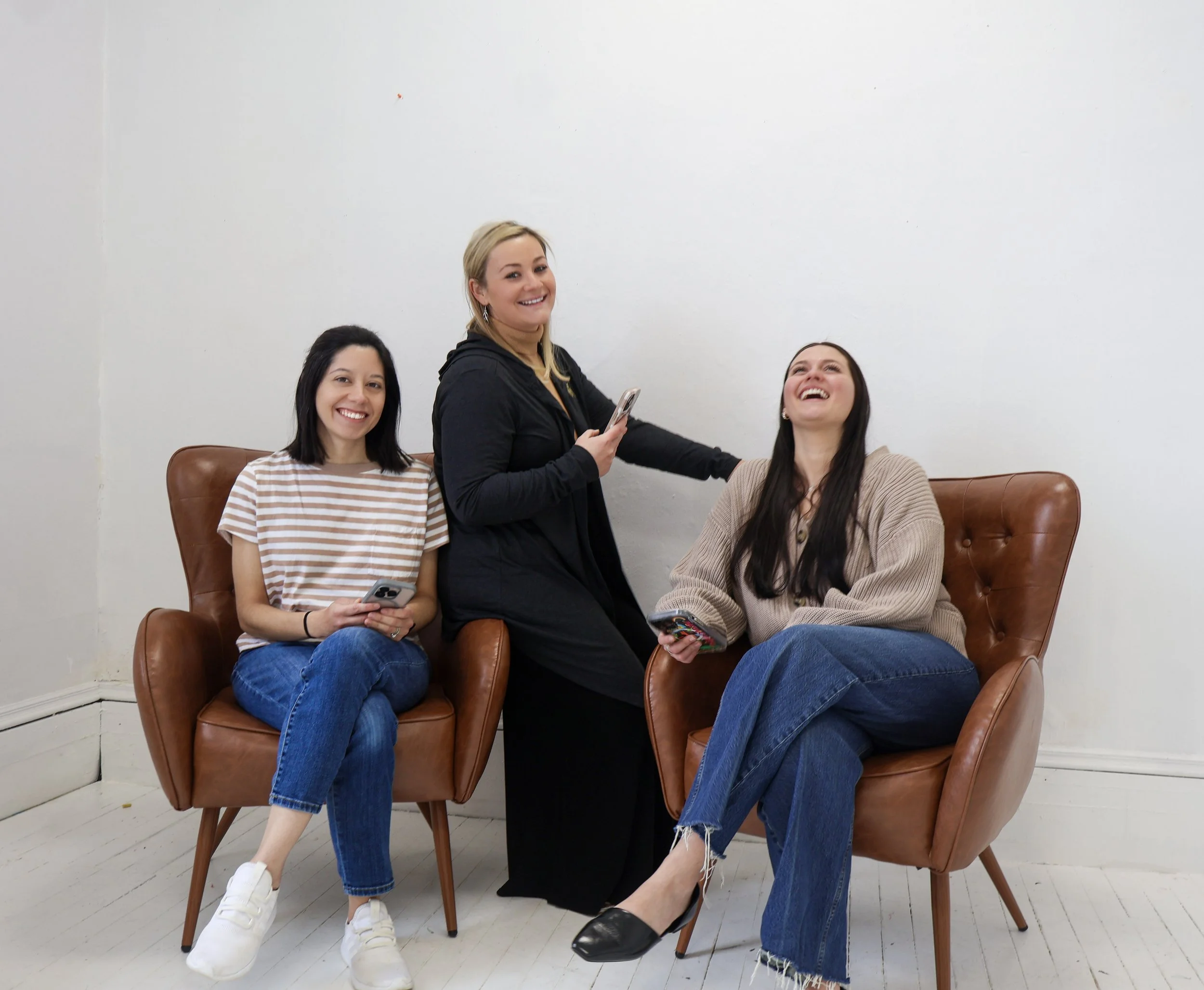 Four women sitting and standing on a brown leather chair against a white wall, smiling and holding smartphones. The woman in the middle stands and holds a phone, the others are seated nearby.