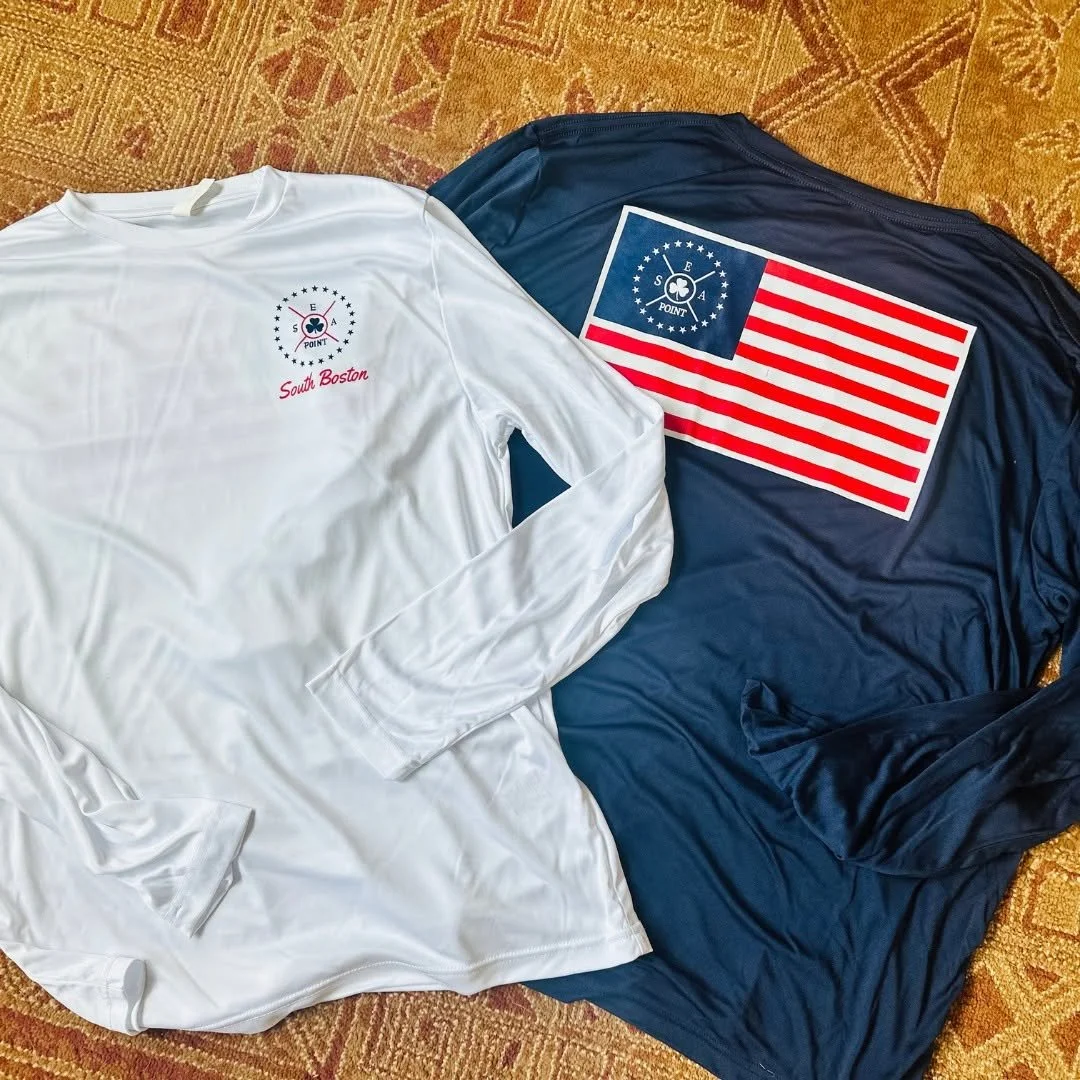 Two long-sleeve shirts, one white with a logo and pop of red text reading 'South Boston,' and one navy with a large American flag and a smaller patriotic logo on the chest, are laid out on a patterned carpet.