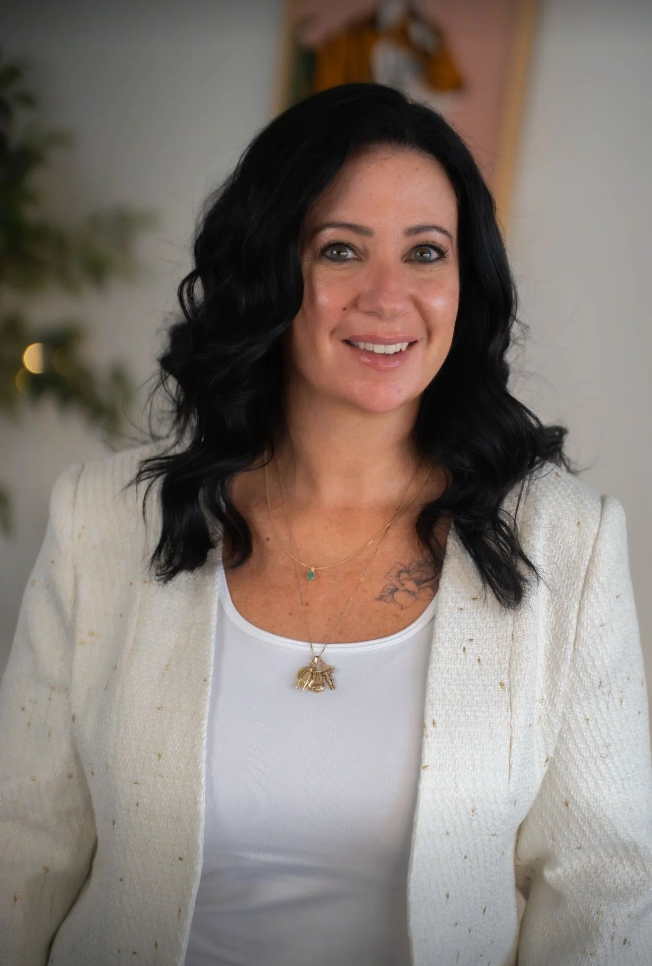A woman with long black wavy hair wearing a white top and a cream-colored blazer, smiling indoors with blurred background.