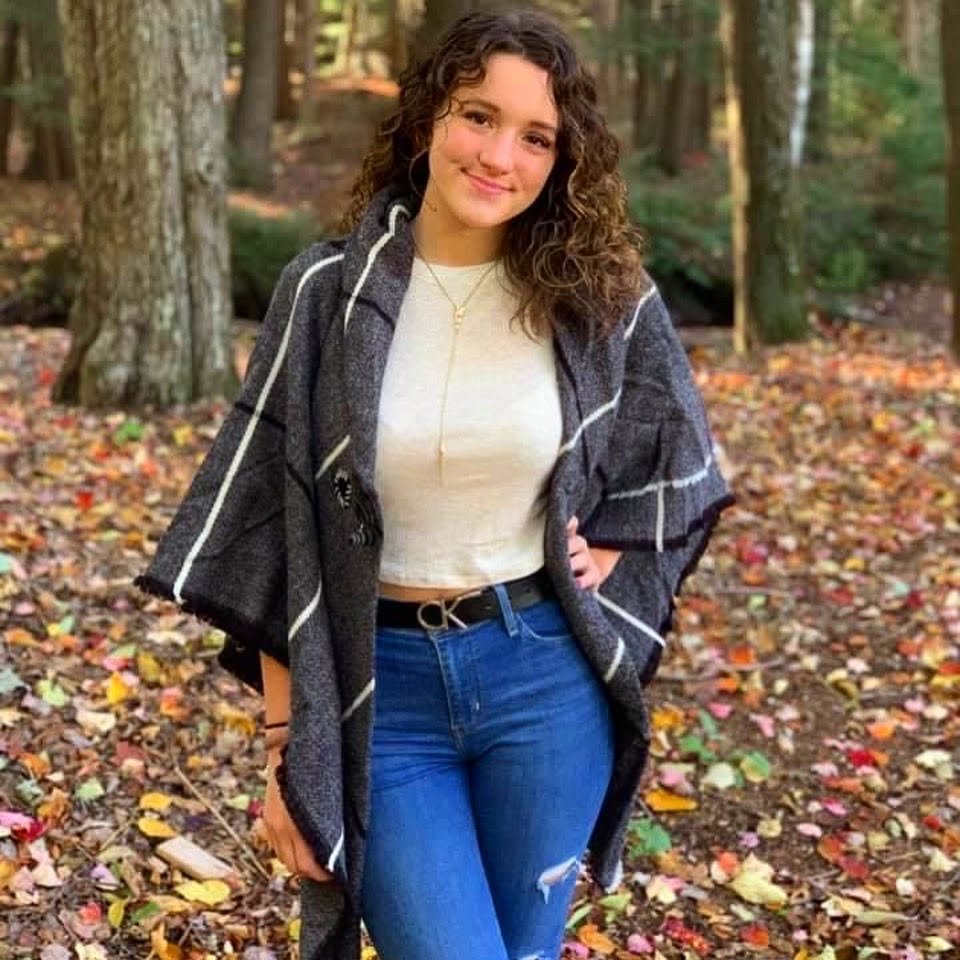 Young woman with curly hair wearing a gray plaid shawl, white top, and blue jeans in an autumn forest